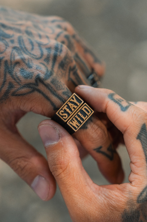 A tattooed hand wears a handcrafted copper Relentless Betrayal Stay Wild Ring engraved with STAY WILD, while another tattooed hand gently touches the ring.