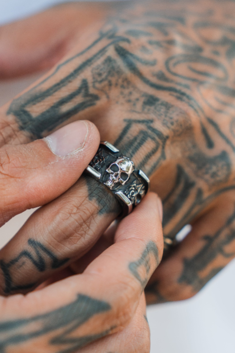 A tattooed hand displays the Relentless Betrayal Outlaw Ring—an intricately detailed silver skull piece, with tattoos visible on the fingers and knuckles.