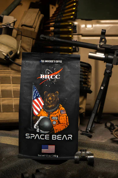 A Space Bear Roast Ground coffee bag from Black Rifle Coffee features a cartoon bear astronaut in an orange suit, holding a helmet and flag. Set against military gear, it offers a light roast with nutty aromas, perfect for adventurous brew seekers.