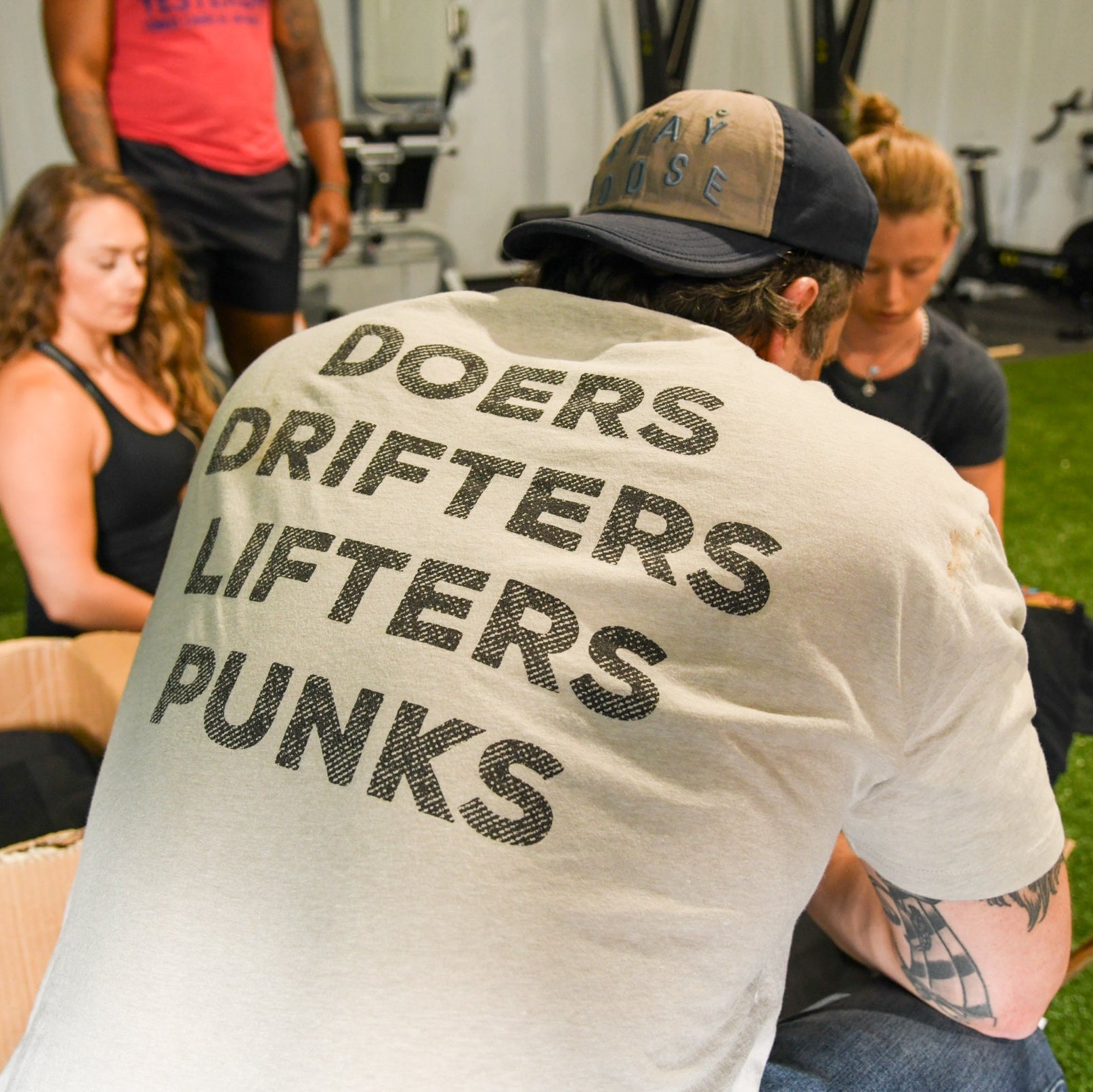 A man in fitness gear wears the Tango Charlie Apparel Doers Drifters Lifters Punks Unisex Tee while sitting indoors with two women; gym equipment is visible. Show off your crew and style at the gym with this bold tee.