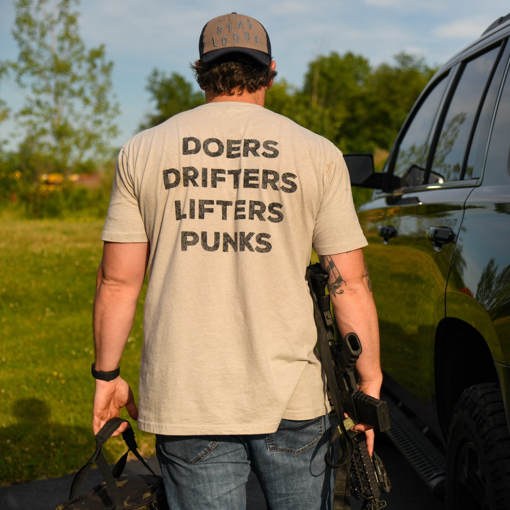 Wearing the Doers Drifters Lifters Punks Unisex Tee by Tango Charlie Apparel, a person stands outdoors by a black SUV with a rifle and bag. Trees and grass frame the scene—fitness gear for those who rep their crew boldly.