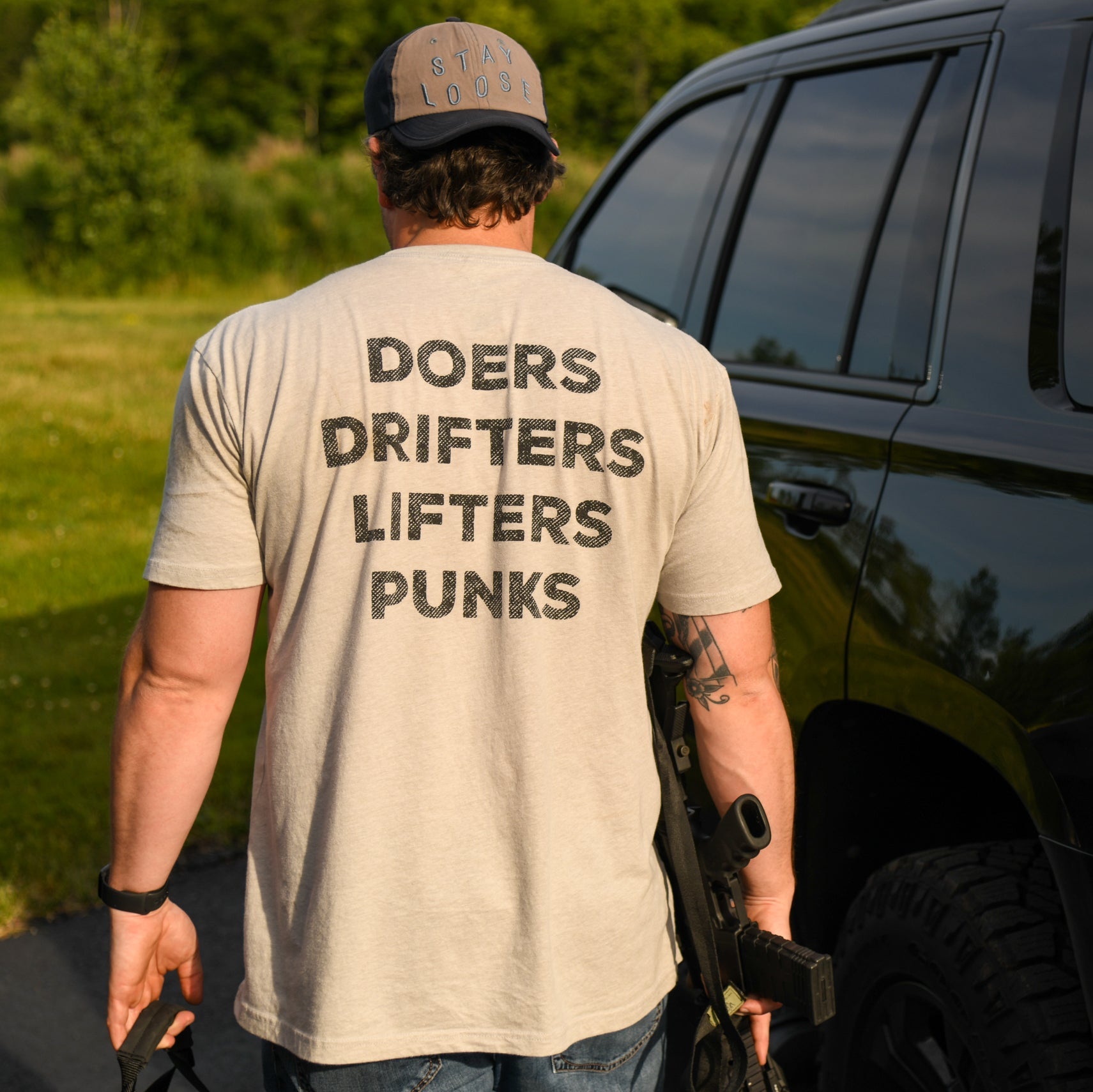 A man wears the Doers Drifters Lifters Punks Unisex Tee by Tango Charlie Apparel, paired with a cap and beige fitness wear, standing outdoors by a black SUV with a rifle—rep your crew in style.