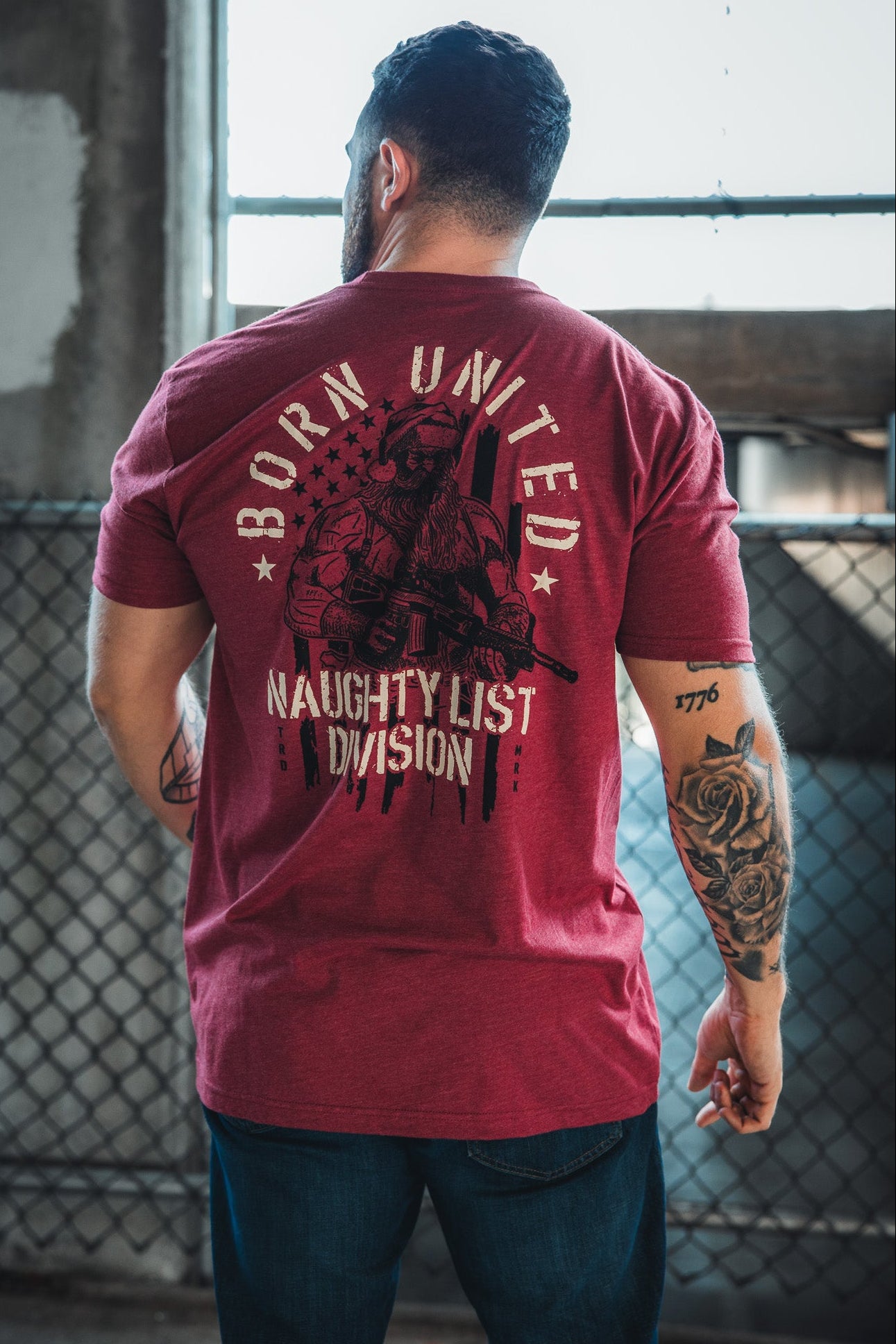 A tattooed man stands indoors by a chain-link fence, wearing the Born United Private Label Tactical Santa Tee—a limited-edition shirt featuring a soldier, American flag, and "BORN UNITED NAUGHTY LIST DIVISION" for bold Christmas spirit.