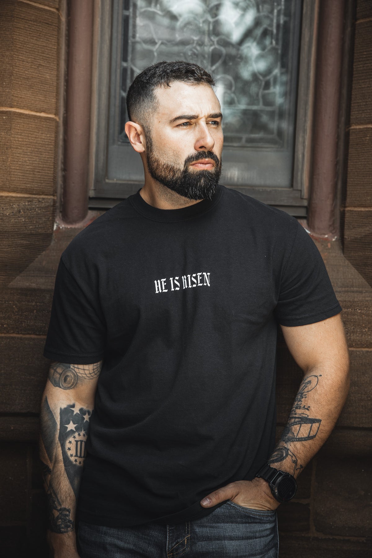 A bearded, tattooed man stands in front of a stone building wearing the 13th "He is Risen Premium Tee," looking to the side with a serious expression.