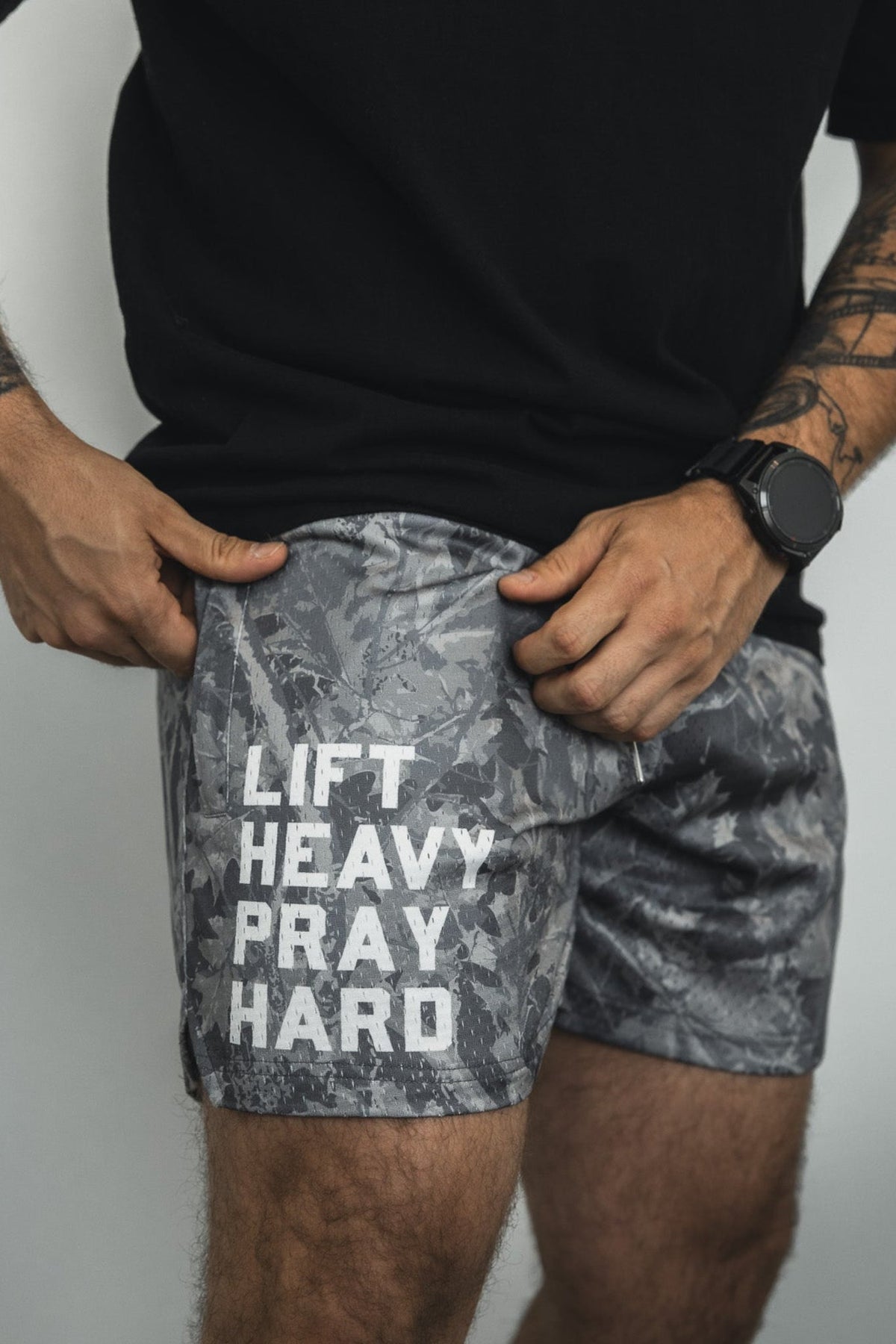 A person wears Tango Charlie Apparel Men's Mesh Lifestyle Shorts (5.5" - Lift Heavy Pray Hard Deadwood), a black t-shirt, and a black wristwatch, with hands resting on the gray patterned shorts.