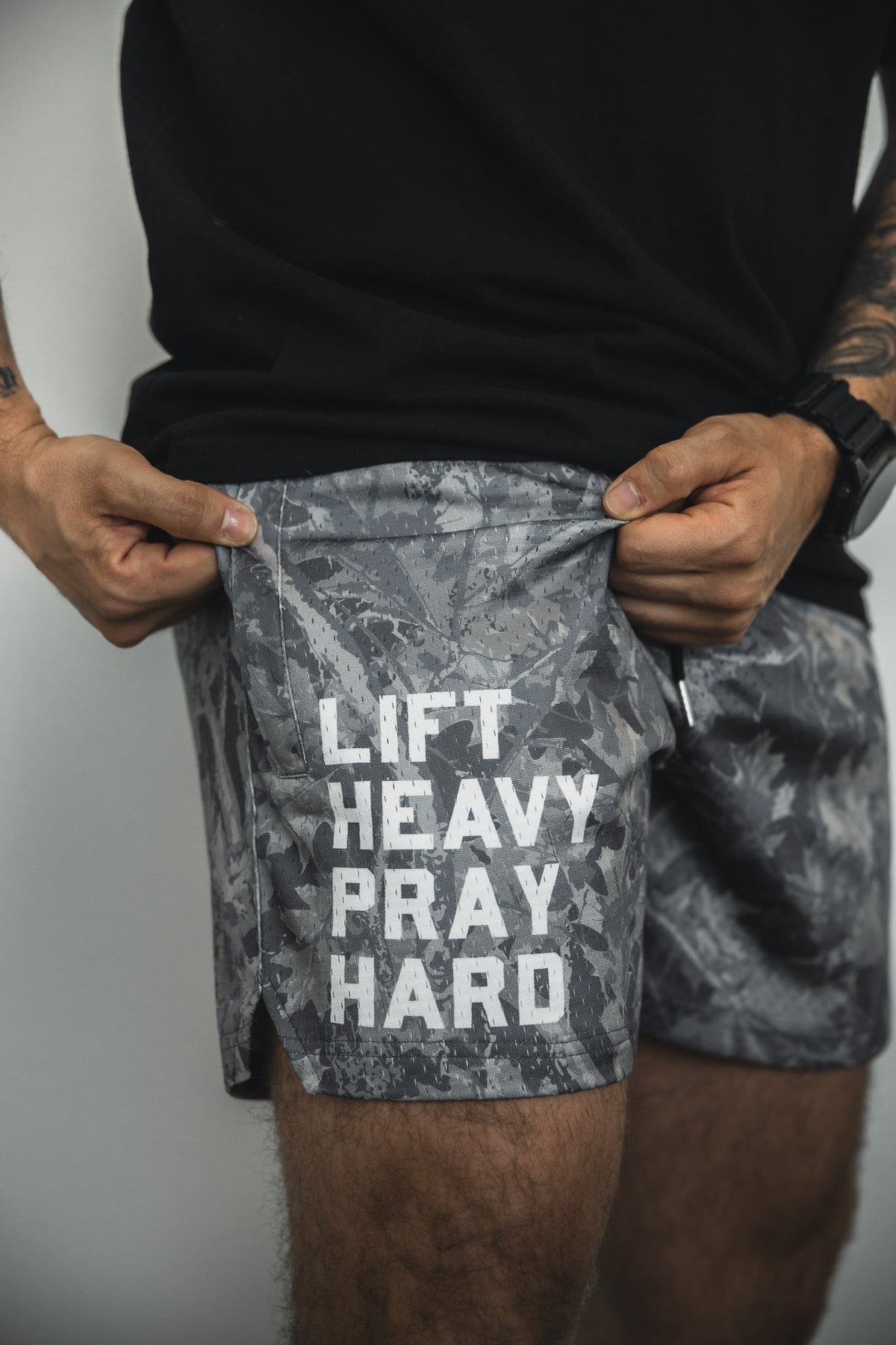 A person wearing Tango Charlie Apparel's Men's Mesh Lifestyle Shorts | 5.5" - Lift Heavy Pray Hard Deadwood in gray camo, pulling the fabric to reveal the "LIFT HEAVY PRAY HARD" print, styled with a black shirt for fitness or everyday wear.