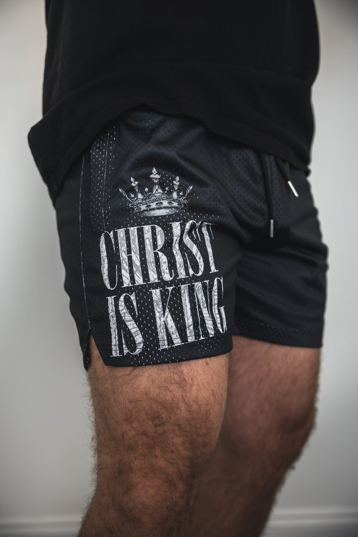 A person wears 13th's Men's Mesh Lifestyle Shorts | 5.5"- Christ is King in black, featuring a crown graphic and text on the left leg, paired with a matching shirt—a standout choice for casual streetwear looks.