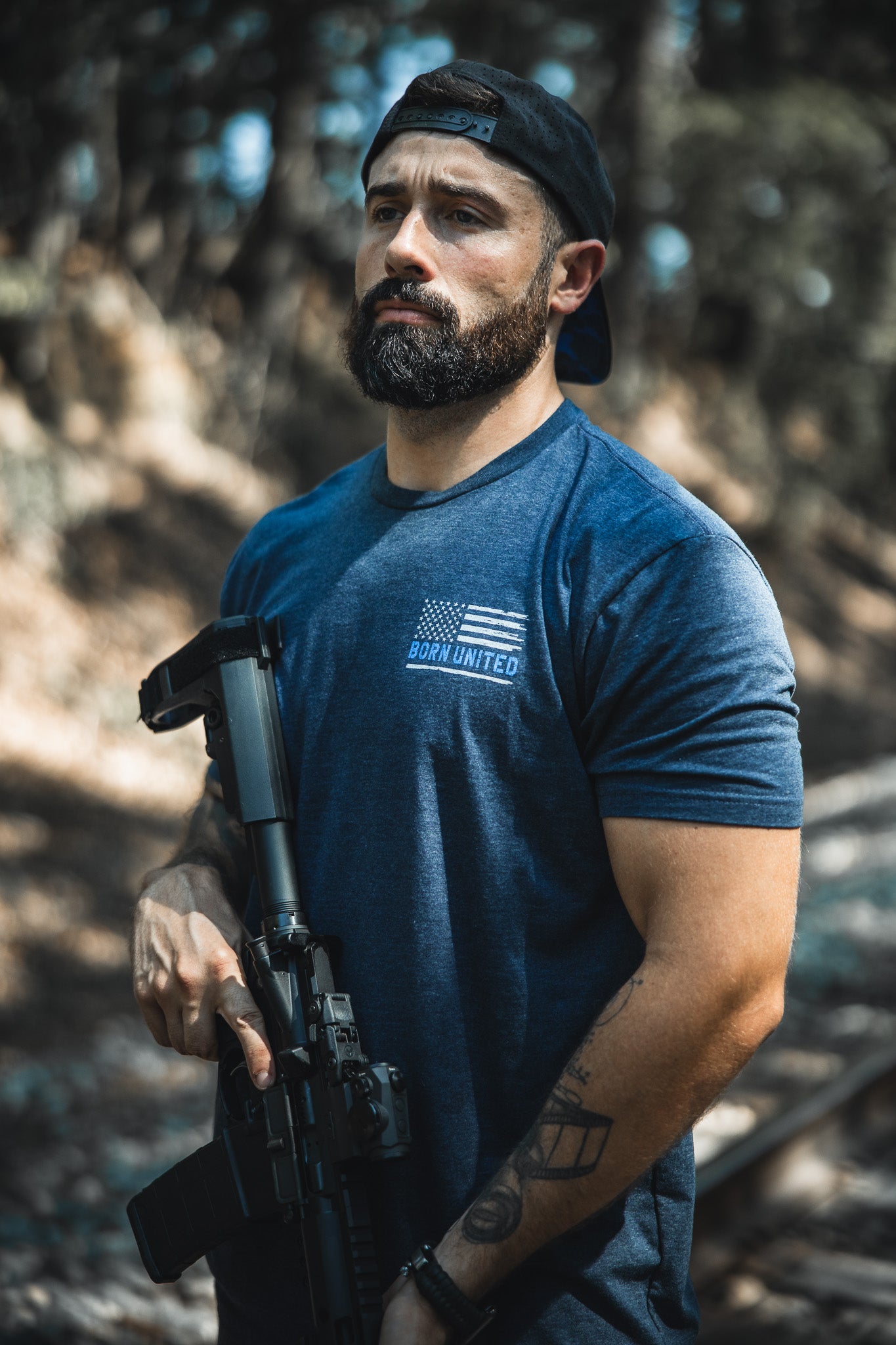 A bearded man in a backwards black cap and a blue The Good Are Unafraid T-shirt by Born United Private Label stands outside, confidently holding a rifle amid trees and sunlight—reflecting bold style and strength.