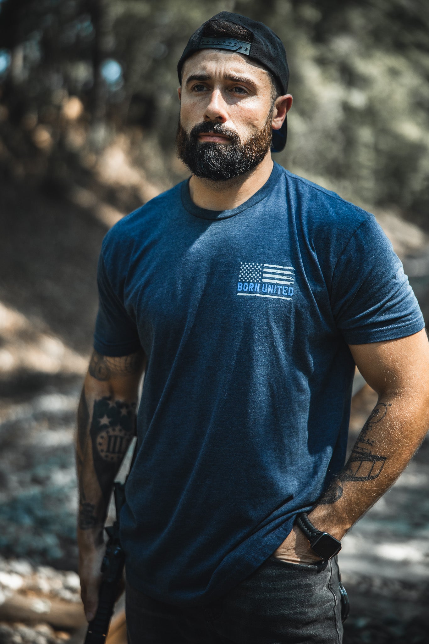 A bearded man in a blue The Good Are Unafraid Born United Private Label tee and a black backward cap stands outdoors with one hand in his pocket, looking ahead seriously. Arm tattoos emphasize the bold style this shirt represents.
