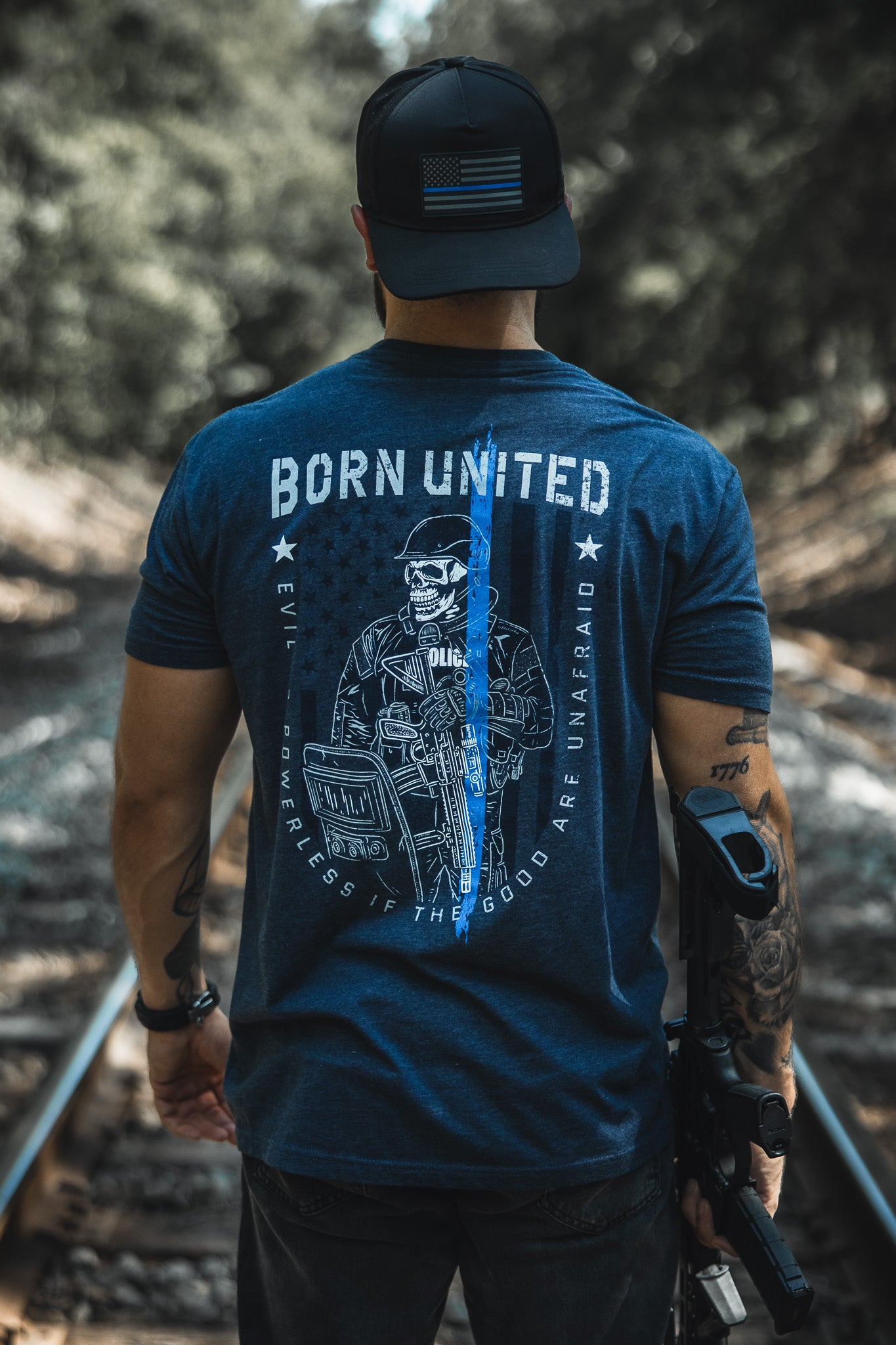 A man stands on railroad tracks wearing a blue The Good Are Unafraid T-shirt by Born United Private Label, featuring a police graphic and blue line. Holding a rifle and wearing an American flag patch cap, he supports Stand Behind the Blue.