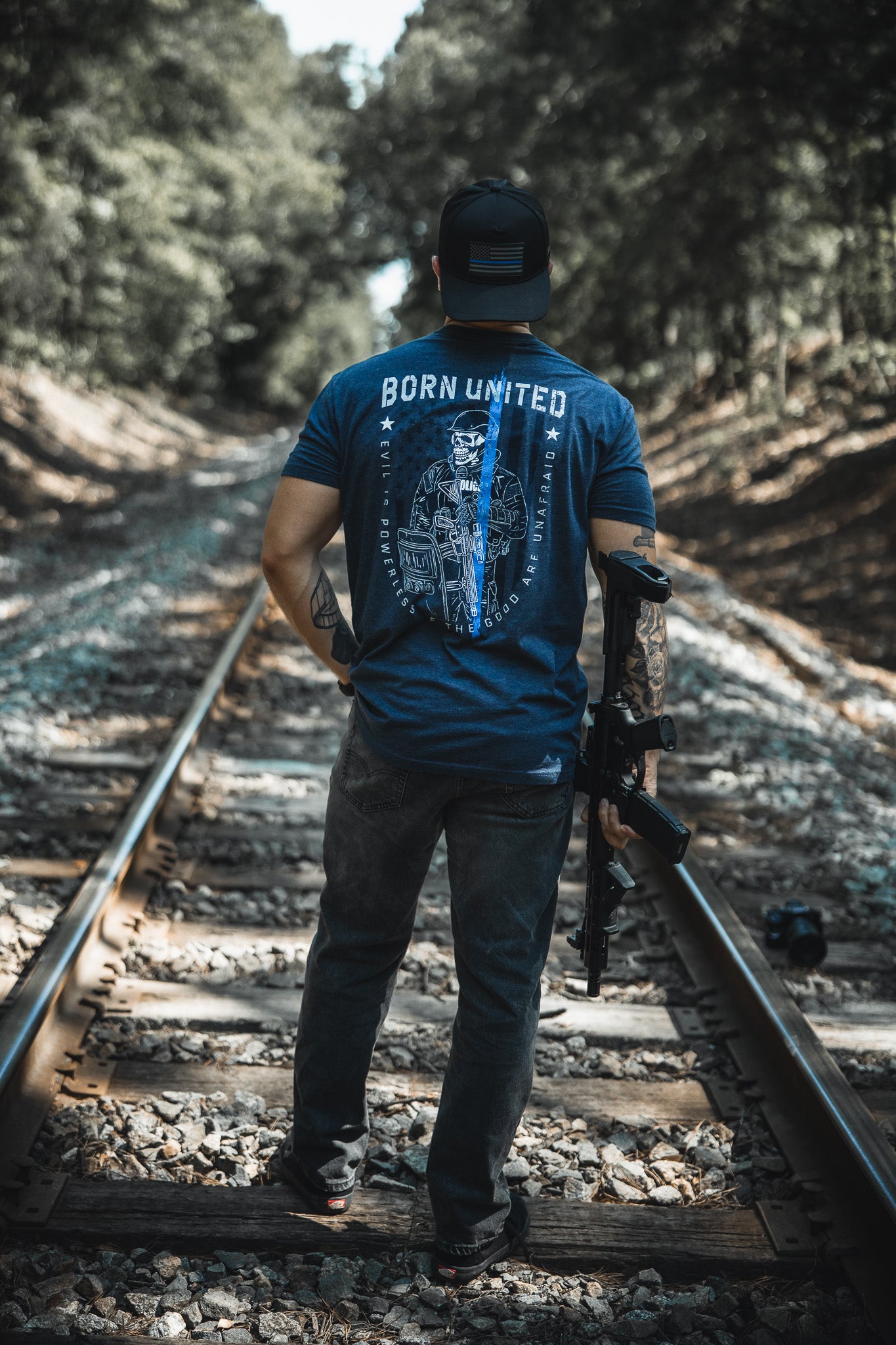A man stands on train tracks, facing away, wearing Born United Private Label’s “The Good Are Unafraid” T-shirt, jeans, and a backwards cap. He holds a rifle in his right hand, with trees lining both sides of the tracks.
