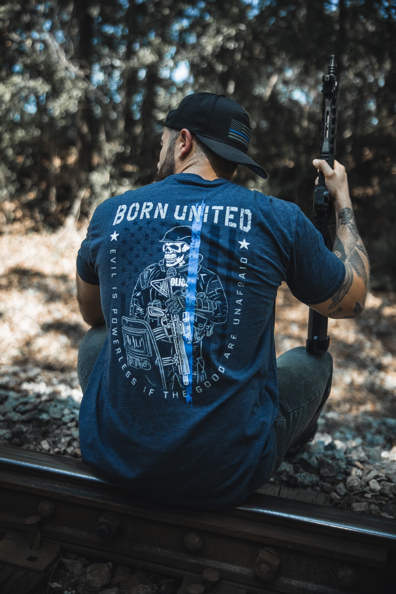 A man wearing the “The Good Are Unafraid” tee by Born United Private Label sits on train tracks in a wooded area, holding a rifle and facing away, embodying the shirt’s message: “Evil is powerless if the good are unafraid.”.