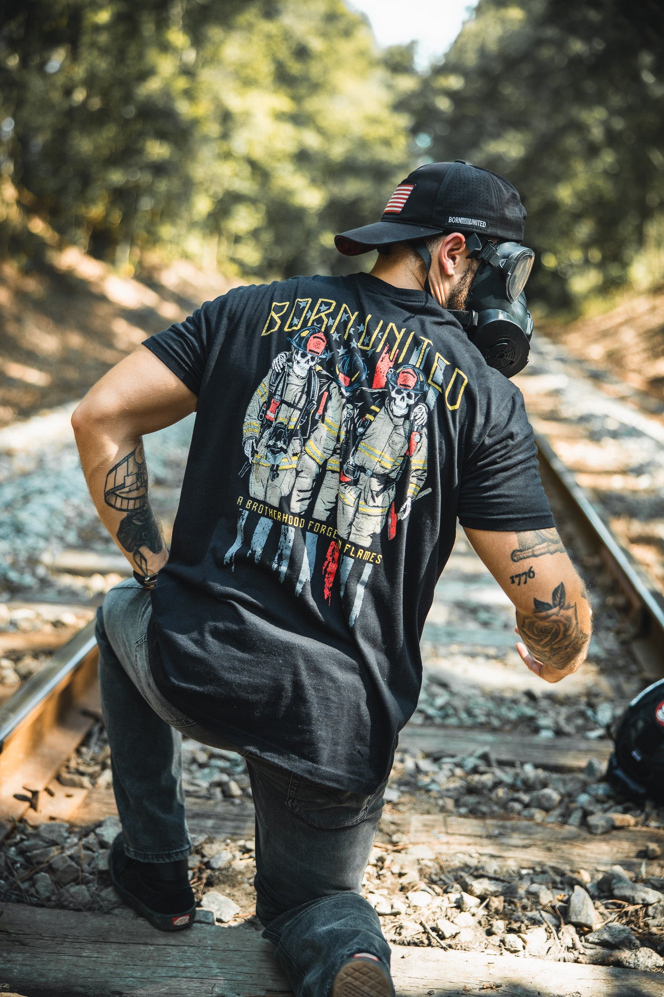 Wearing the Brotherhood Forged In Flames graphic t-shirt by Born United Private Label, a tattooed person in a gas mask and cap kneels on railroad tracks in the woods—reminding us of safetys importance in unconventional settings.