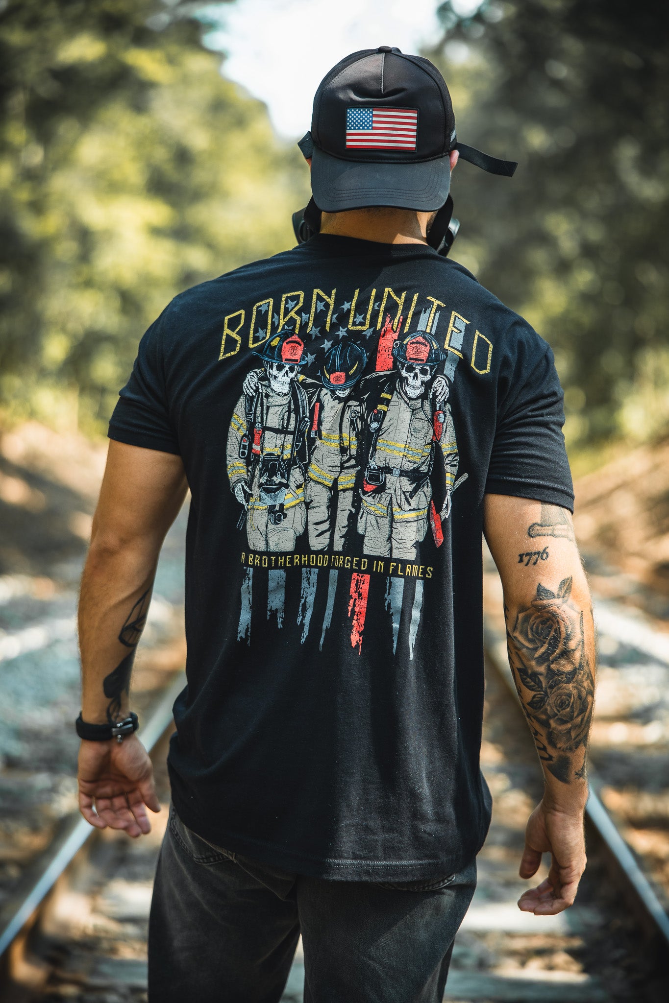 A man in a black Born United Private Label Brotherhood Forged In Flames shirt, showing three firefighters and “BORN UNITED,” and a U.S. flag patch cap stands on train tracks in the woods, his tattooed arm visible from behind.