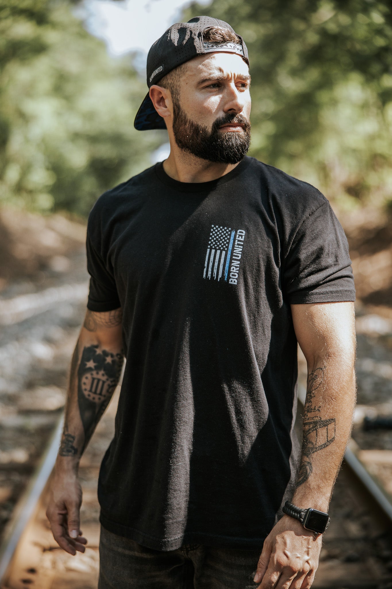 A bearded man in a black backward cap and Born United Private Labels Blue Line Flag Tee stands on railroad tracks outdoors, looking to the side, showing support for law enforcement amid trees and sunlight.
