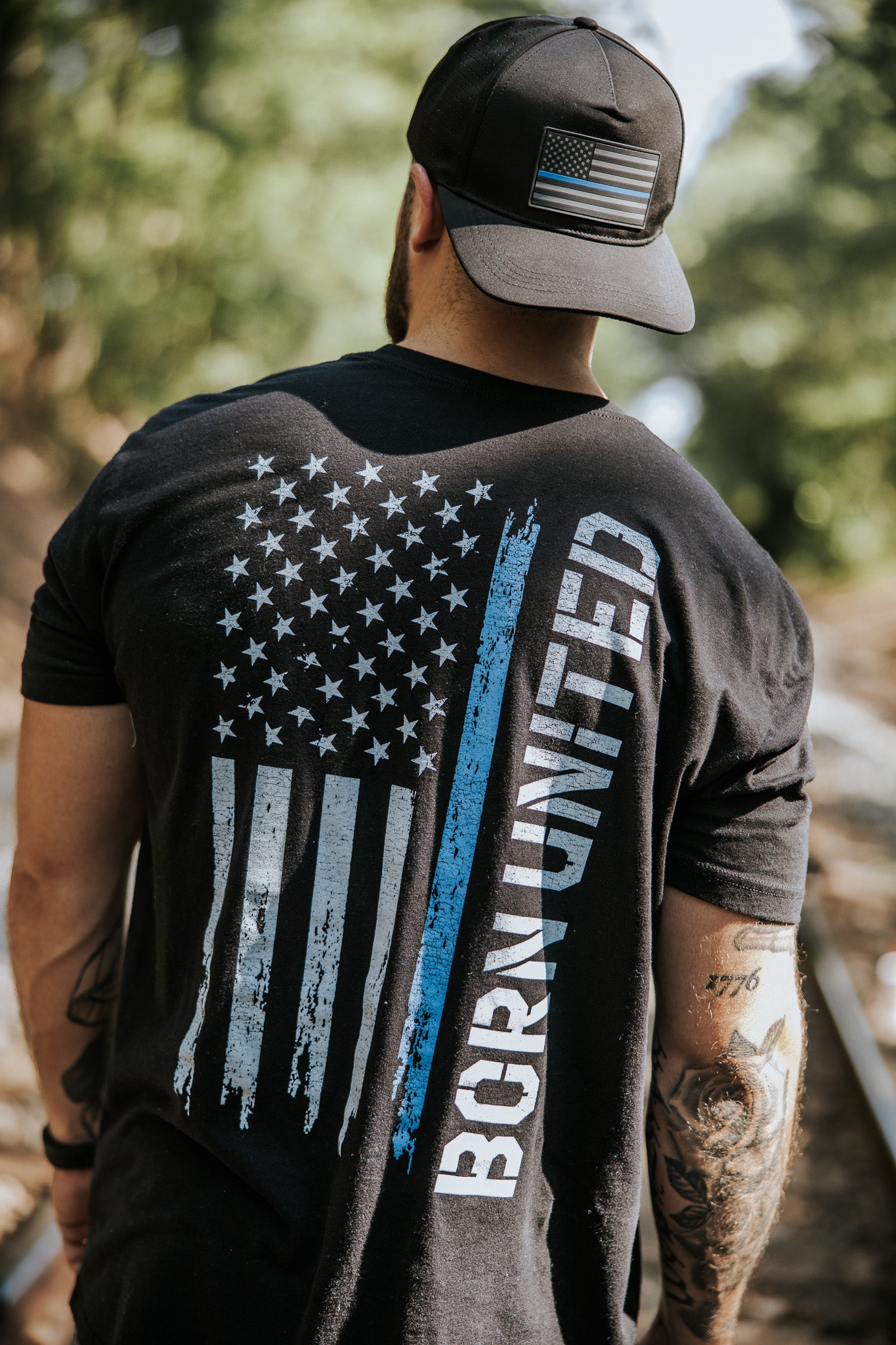 A man stands outdoors on a sunlit path, wearing the Born United Private Label Blue Line Flag Tee—a black T-shirt featuring a blue and white American flag and showing support for Back the Blue.