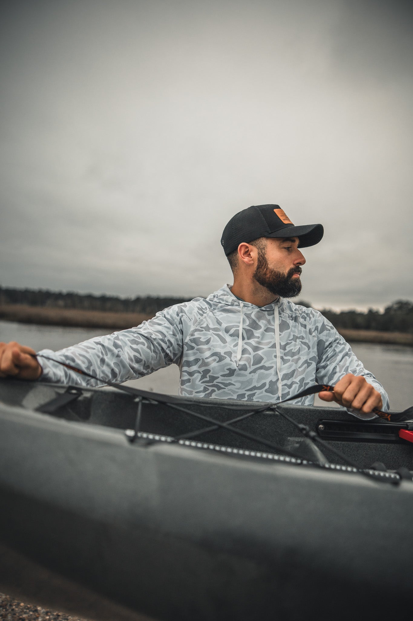 A bearded man in a black cap and Mucker Bucket Performance Hoodie - Ghost Camo sits in a kayak on calm water, looking left, with an overcast sky and shoreline visible in the background.