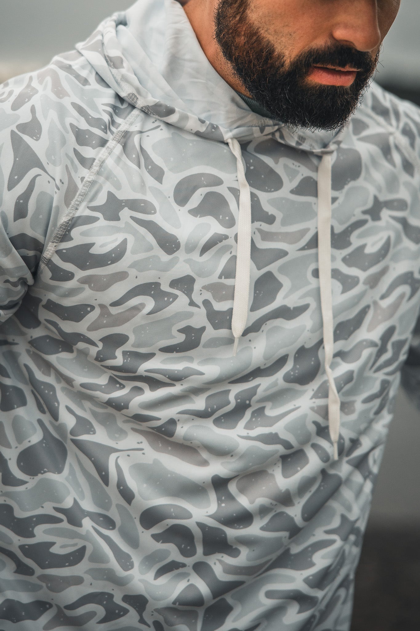 A man with a dark beard models the Mucker Bucket Performance Hoodie - Ghost Camo, featuring light gray and white camouflage with white drawstrings. The cropped image shows his upper torso and part of his face.