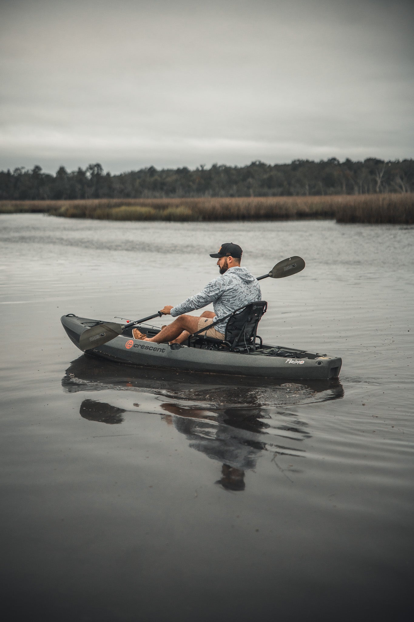 Wearing a Mucker Bucket Performance Hoodie - Ghost Camo and a cap, a person paddles a gray kayak on a calm river with grassy banks and trees under an overcast sky.