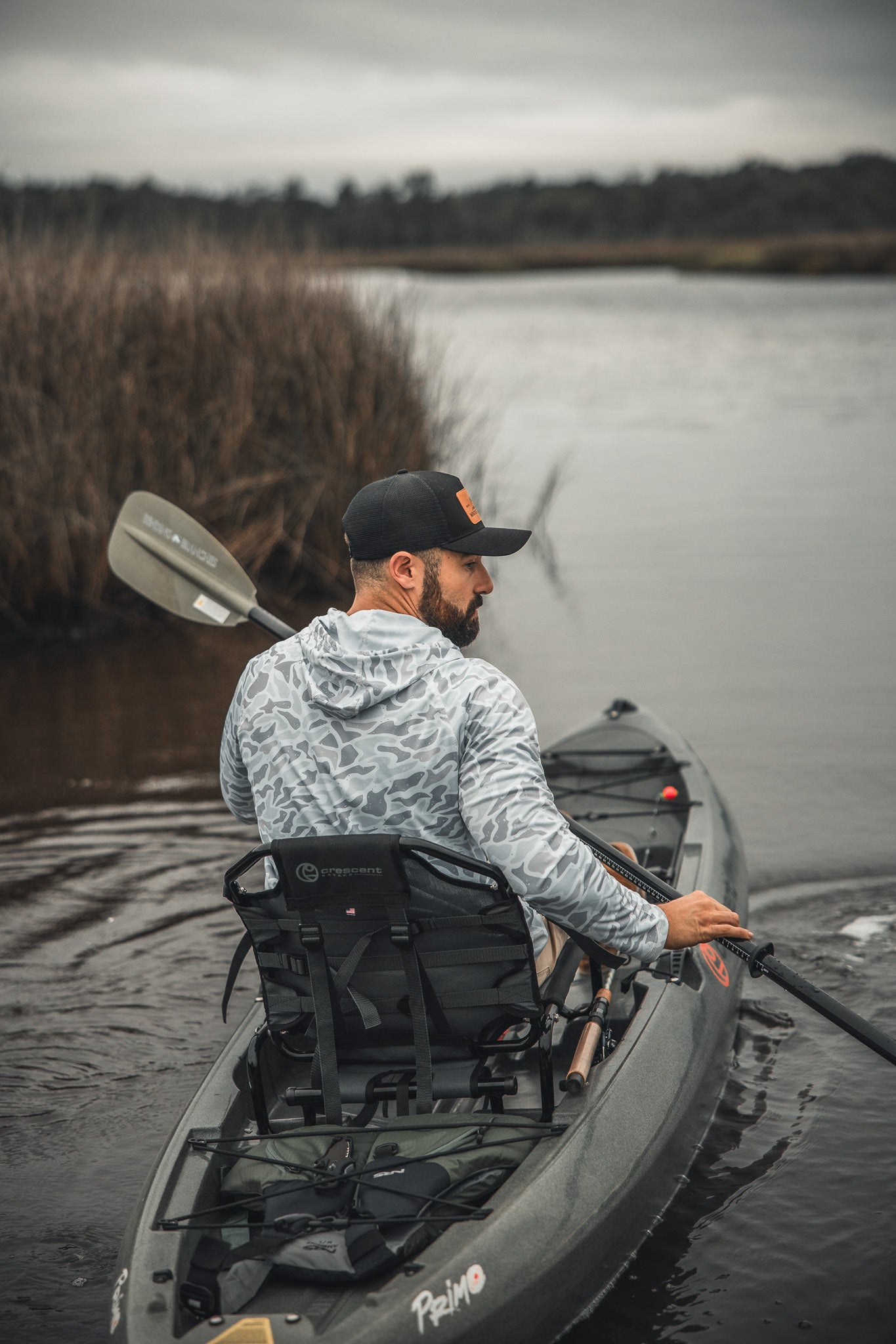 A man wearing a Mucker Bucket Performance Hoodie - Ghost Camo and a black cap paddles his kayak down a calm, overcast river bordered by reeds and grassy marshland.