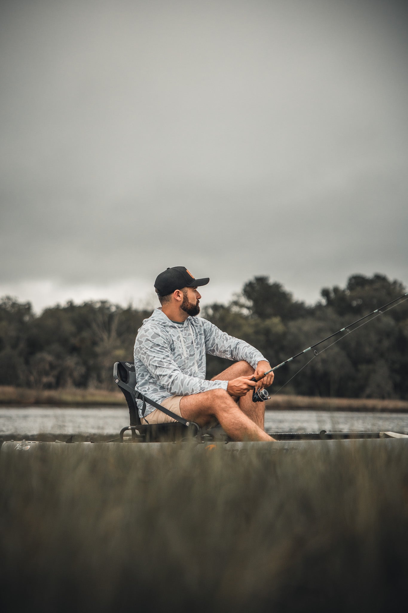 A man in the Mucker Bucket Performance Hoodie - Ghost Camo sits on a folding chair by the water, fishing rod in hand, with trees and a cloudy sky behind him.