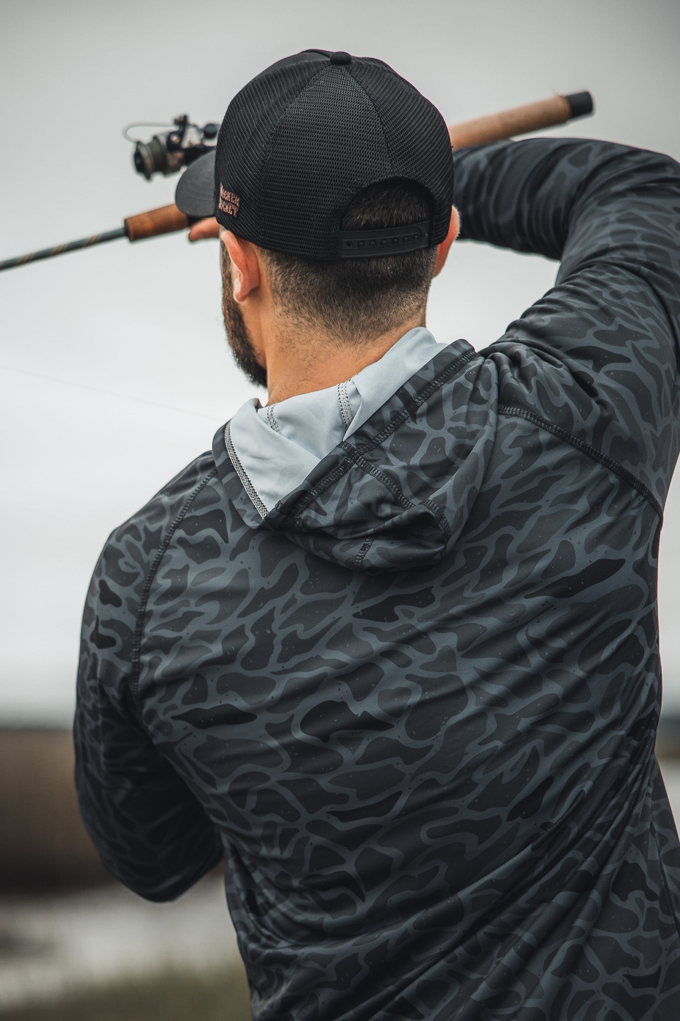 A man wearing the Mucker Bucket Performance Hoodie - Stealth Camo faces away from the camera, fishing with a rod over his shoulder.