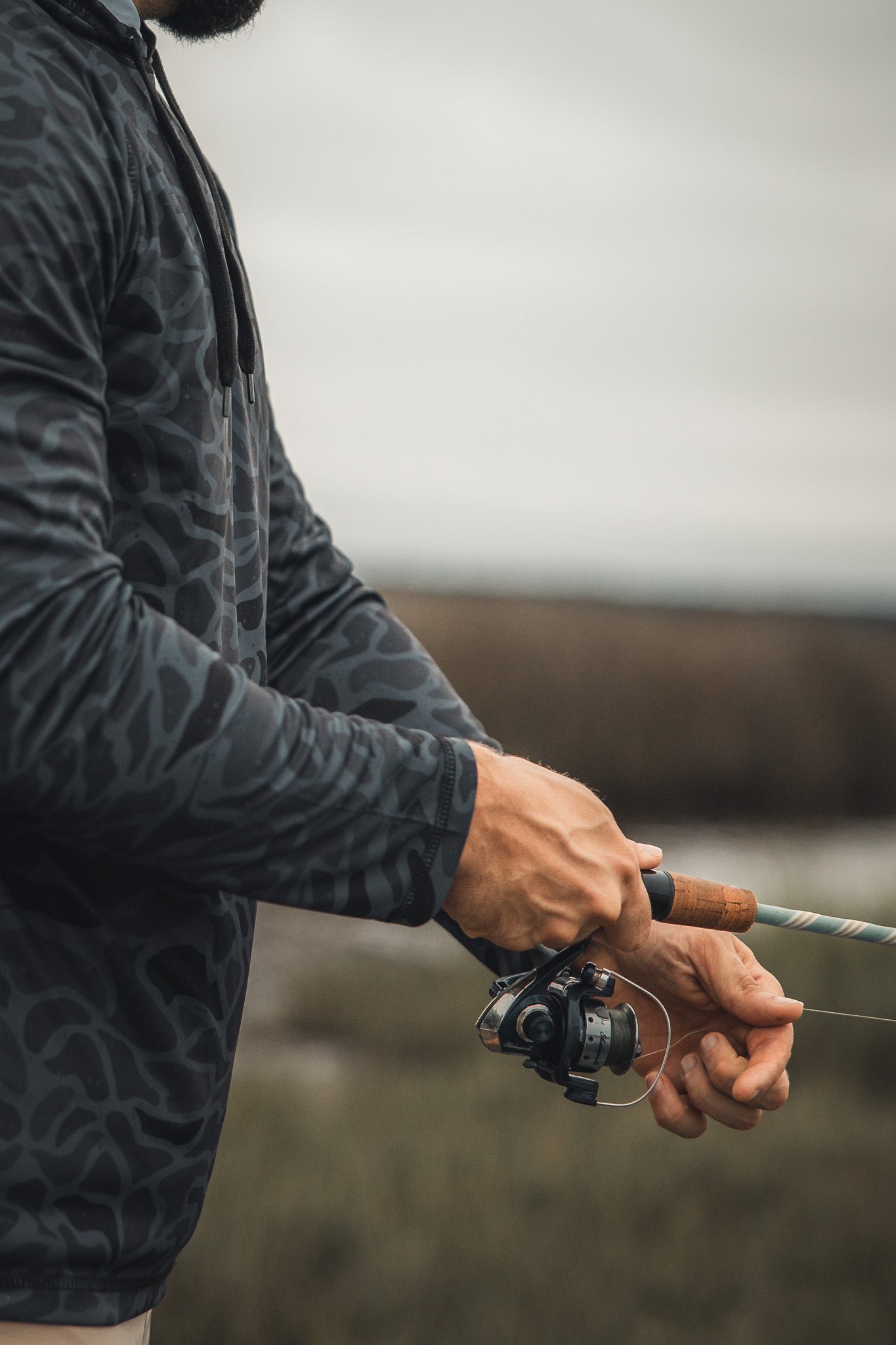 A person in a Mucker Bucket Performance Hoodie - Stealth Camo holds a fishing rod and reel, ready to cast outdoors on an overcast day. Only their upper body and hands are visible.