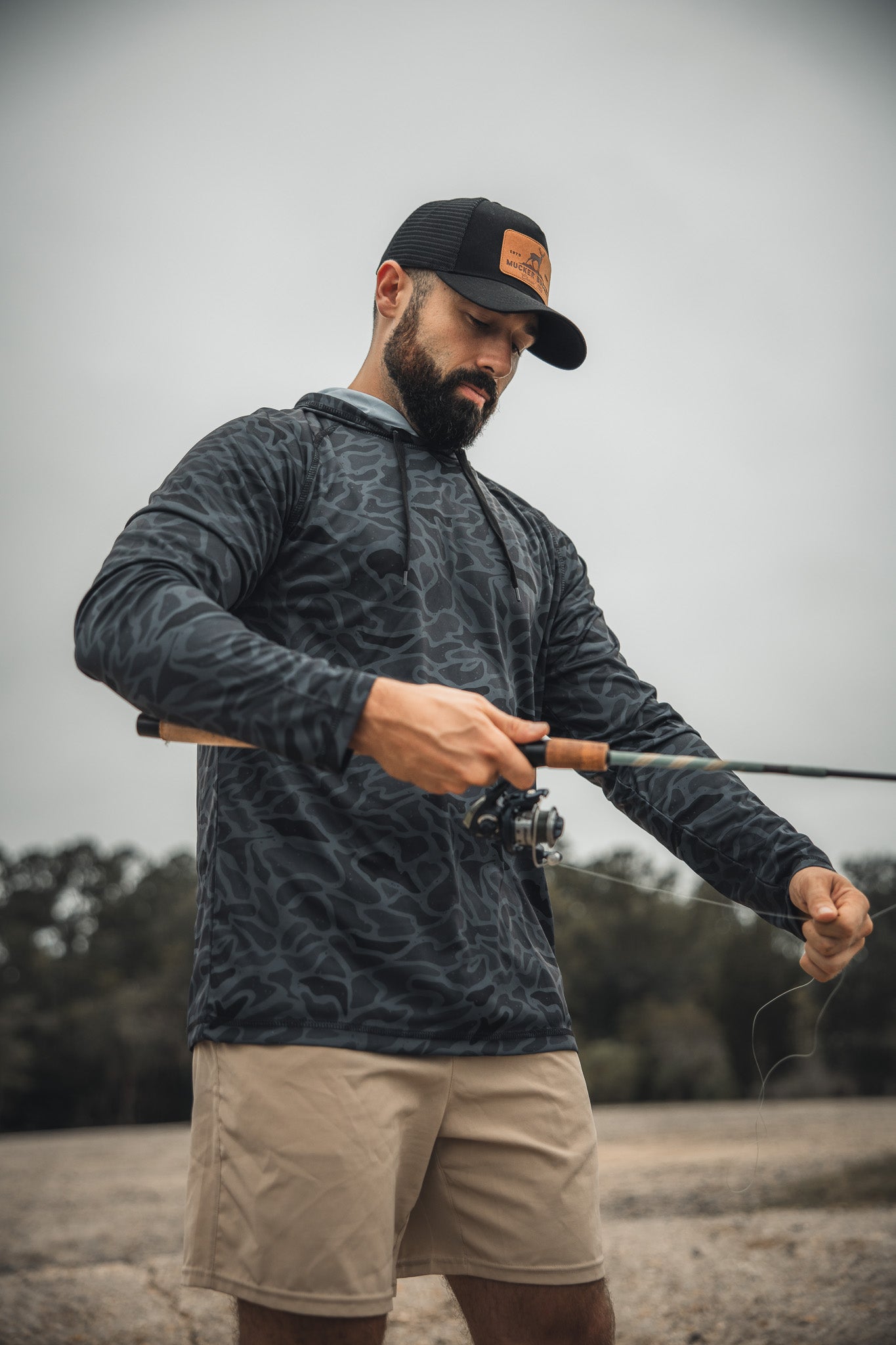 A man wearing the Mucker Bucket Performance Hoodie - Stealth Camo, a black hat, and beige shorts is outdoors with a fishing rod, adjusting the line against a backdrop of trees and a cloudy sky.