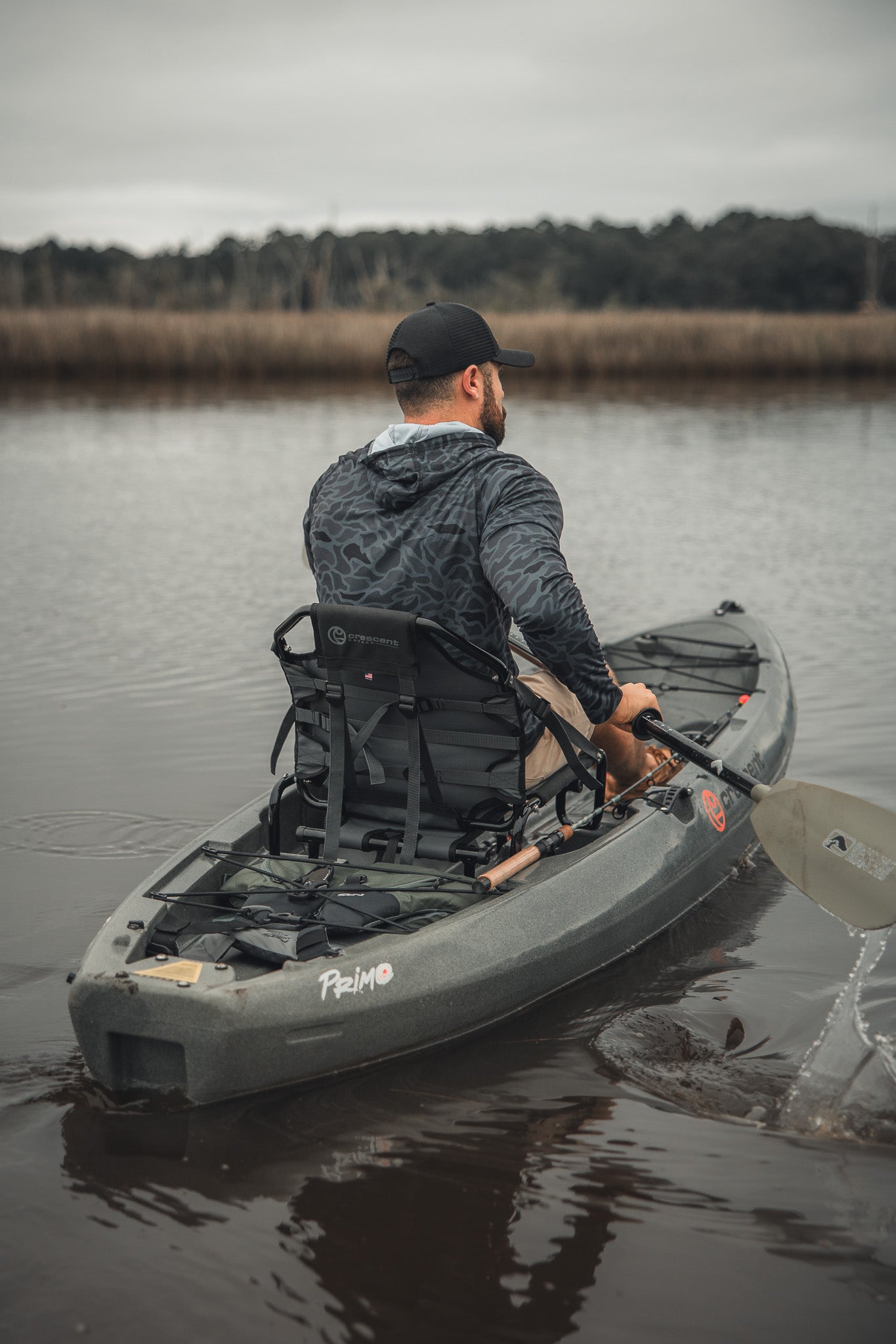 A man wearing a Mucker Bucket Performance Hoodie - Stealth Camo and a black cap paddles a gray kayak on calm water, surrounded by tall grass and trees under a cloudy sky.