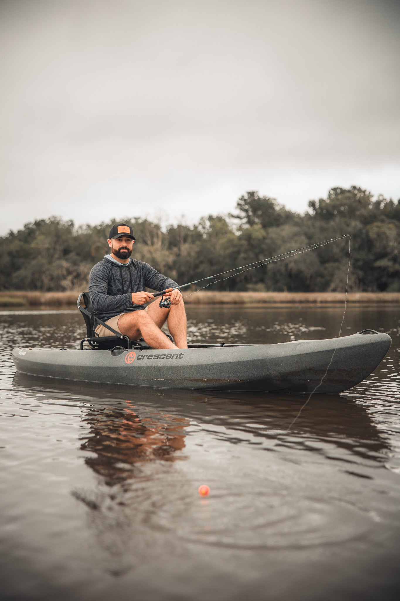 A man in a Mucker Bucket Performance Hoodie - Stealth Camo, sunglasses, and hat fishes from a kayak on a calm, cloudy lake. He holds a rod with the bobber nearby as trees line the background.