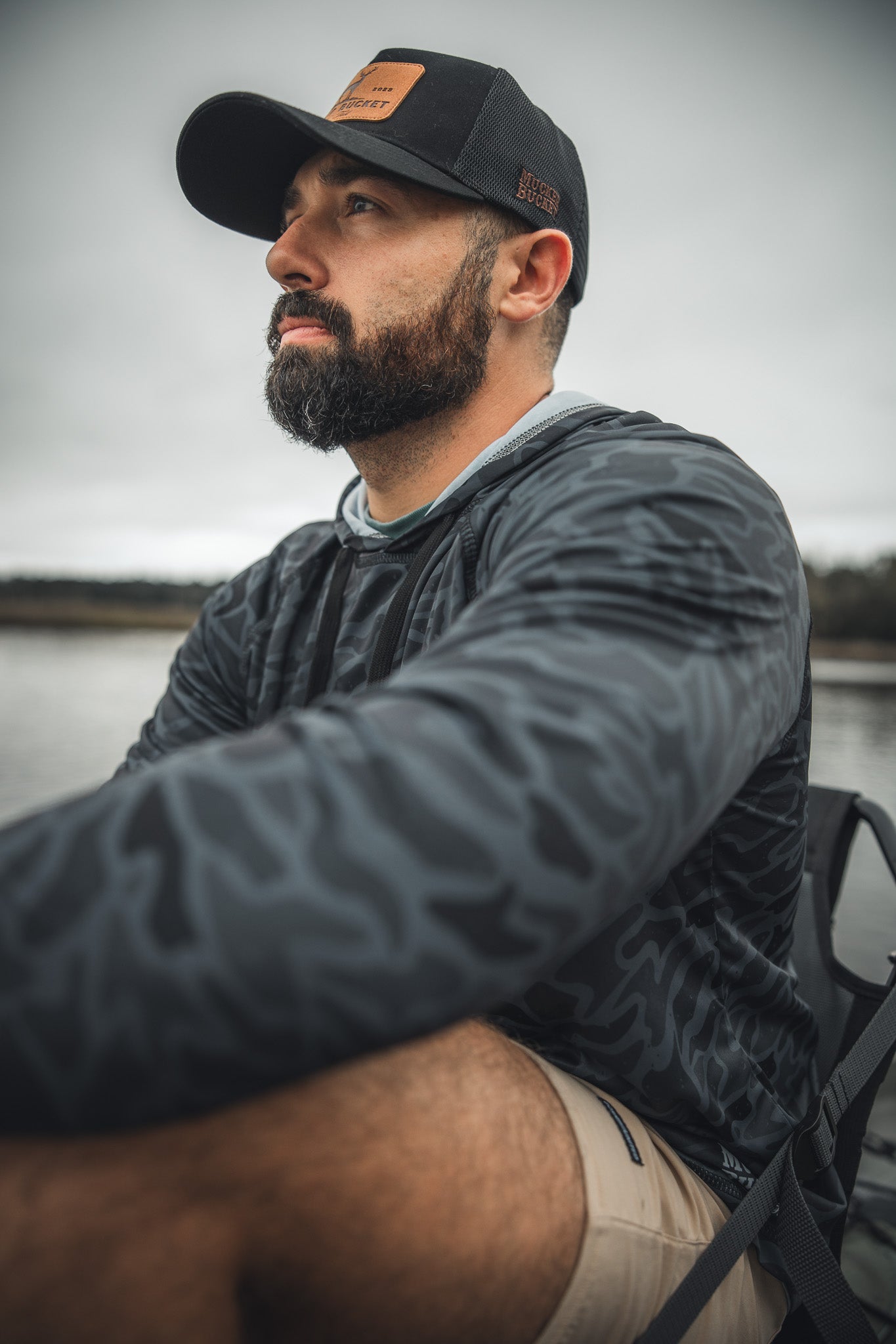 A bearded man in a black cap and Mucker Bucket Performance Hoodie - Stealth Camo sits outdoors in shorts, looking thoughtful. The blurred background hints hes near water on a cloudy day.
