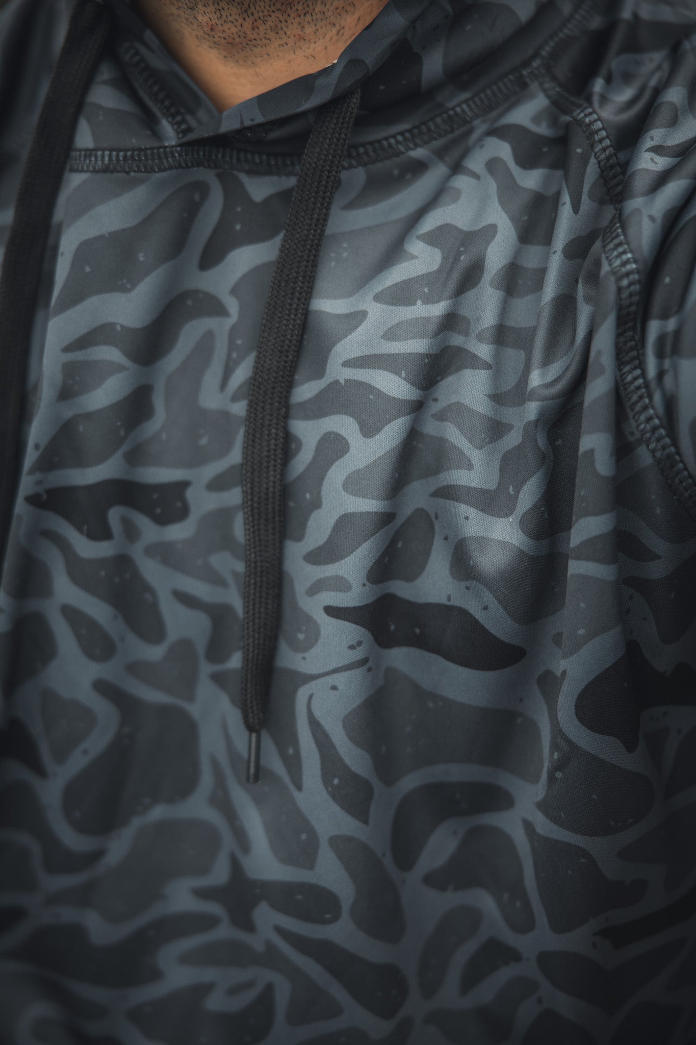 Close-up of a person wearing the Mucker Bucket Performance Hoodie - Stealth Camo, showing an abstract camo pattern on the neck, shoulder, and chest to highlight the athletic fit and lightweight, sleek design.