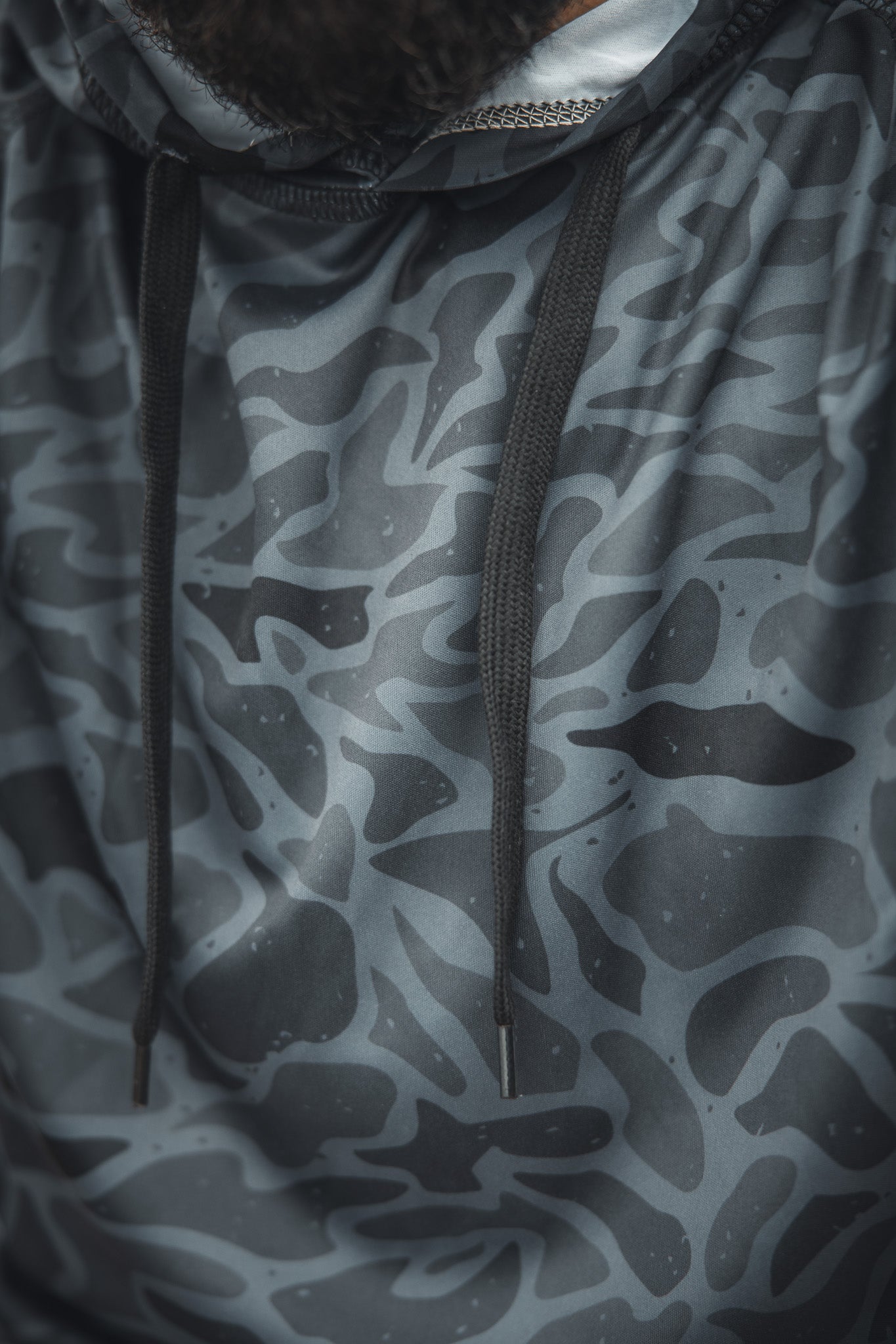 Close-up of the Mucker Bucket Performance Hoodie - Stealth Camo, featuring a gray and black abstract camo pattern with black drawstrings. A portion of a persons beard is visible at the top of the image.