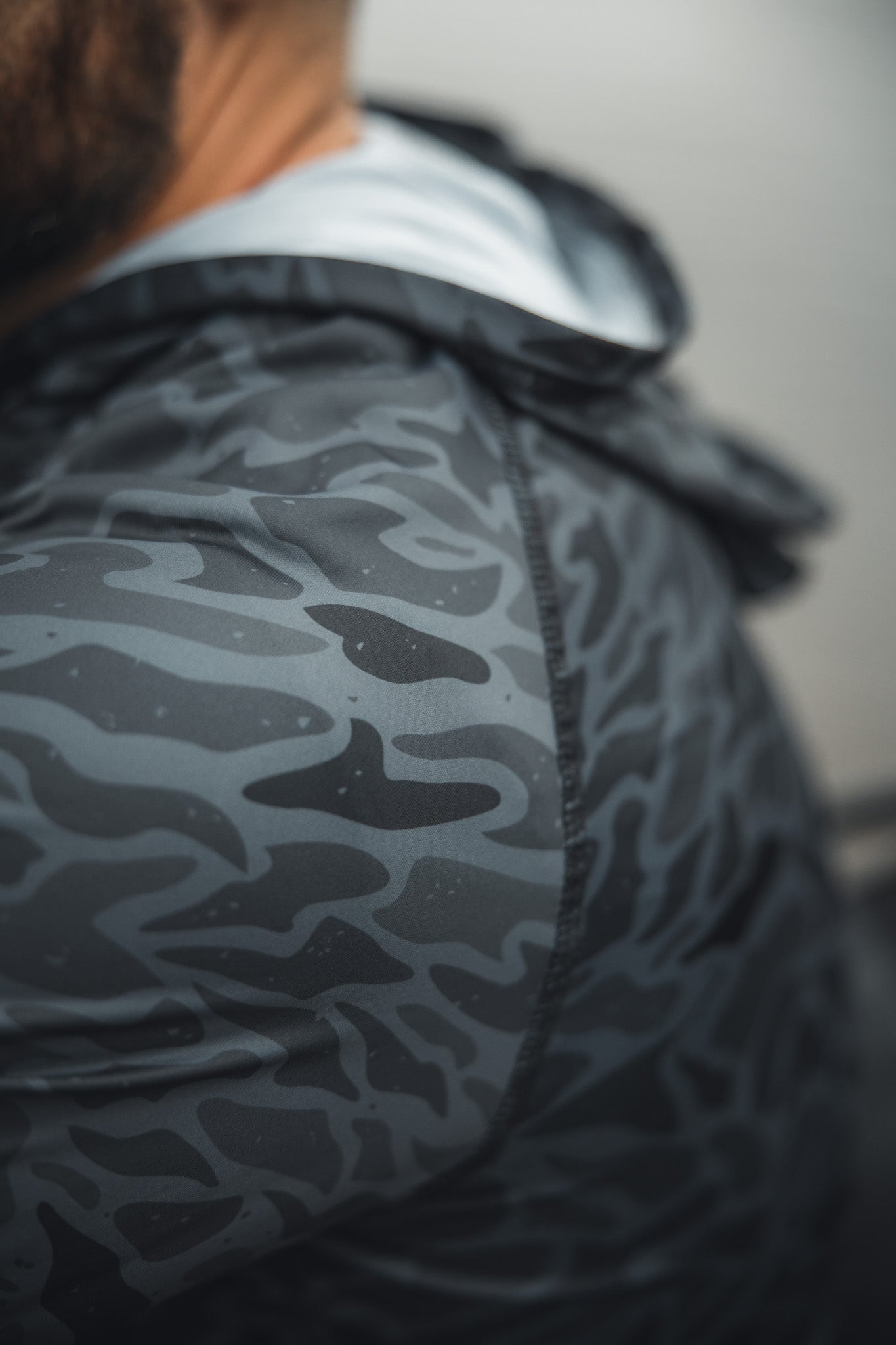 Close-up of a person wearing the Mucker Bucket Performance Hoodie - Stealth Camo, focusing on the shoulder and upper back to highlight its athletic, lightweight design, with part of their neck and beard visible.