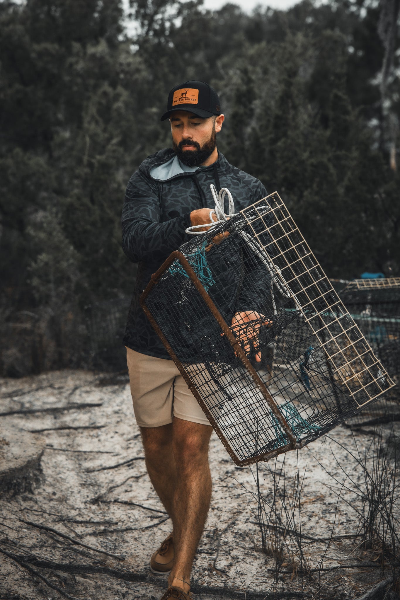 A man wears the Mucker Bucket Performance Hoodie - Stealth Camo, a dark jacket, and tan shorts as he carries a metal cage trap outdoors among trees and rough terrain.