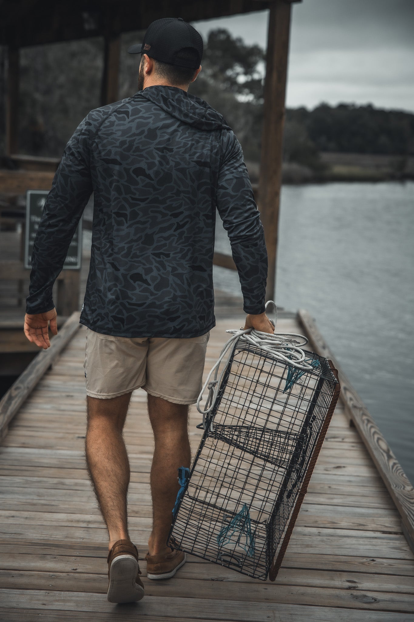A man in a black cap, patterned jacket, and khaki shorts walks on a wooden dock by calm water, carrying an empty wire crab trap while wearing the Mucker Bucket Performance Hoodie - Stealth Camo for changing weather.