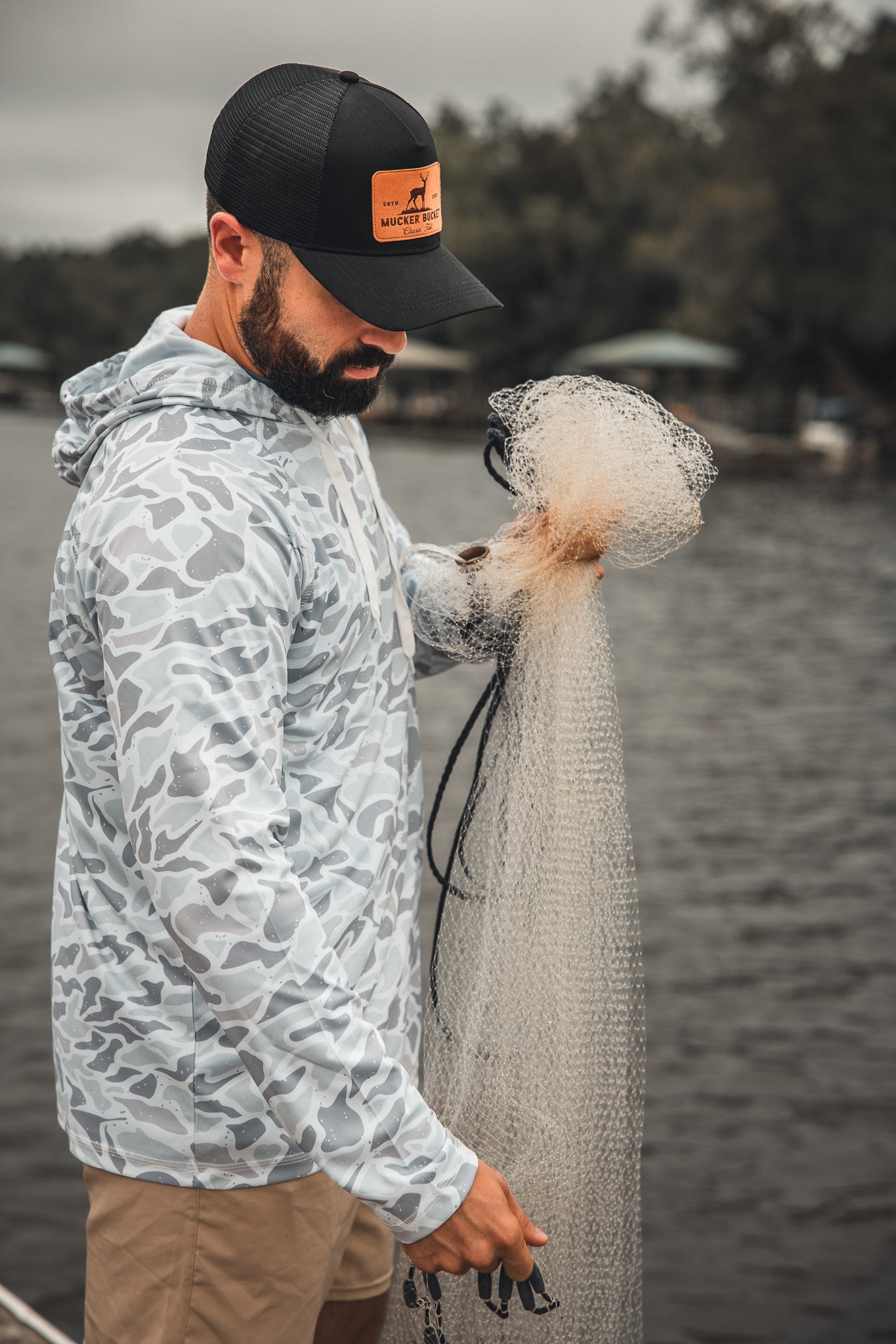A man in the Mucker Bucket Performance Hoodie - Ghost Camo and a black cap stands by the water, untangling a fishing net with trees and houses blurred in the background.