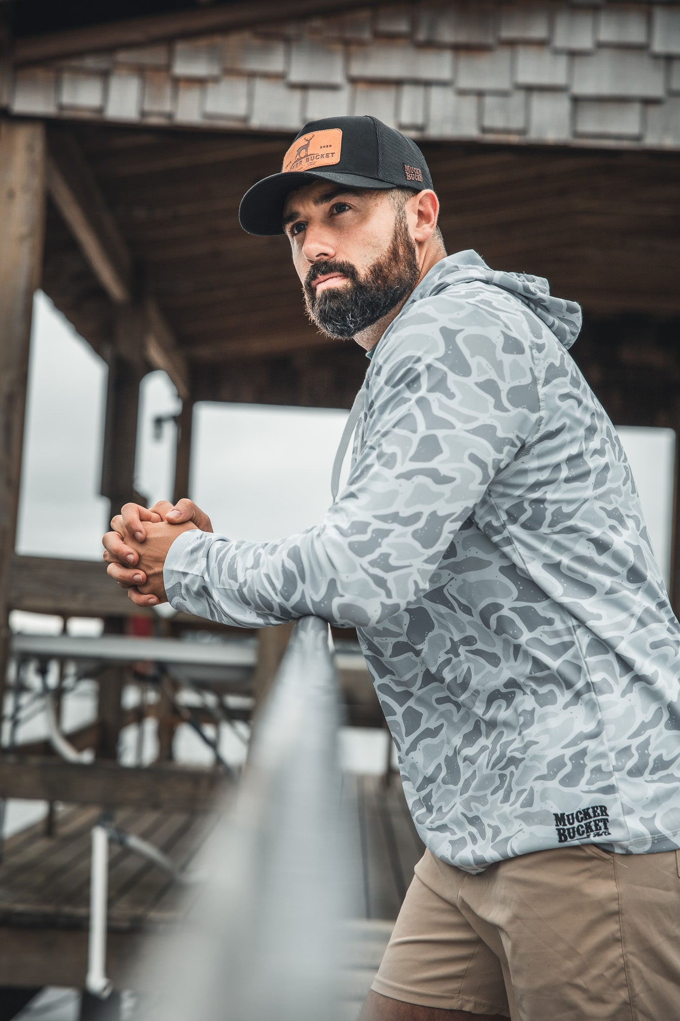 A bearded man in a Mucker Bucket Performance Hoodie - Ghost Camo and a black cap leans on a railing outdoors, gazing thoughtfully into the distance beneath a wooden shelter.