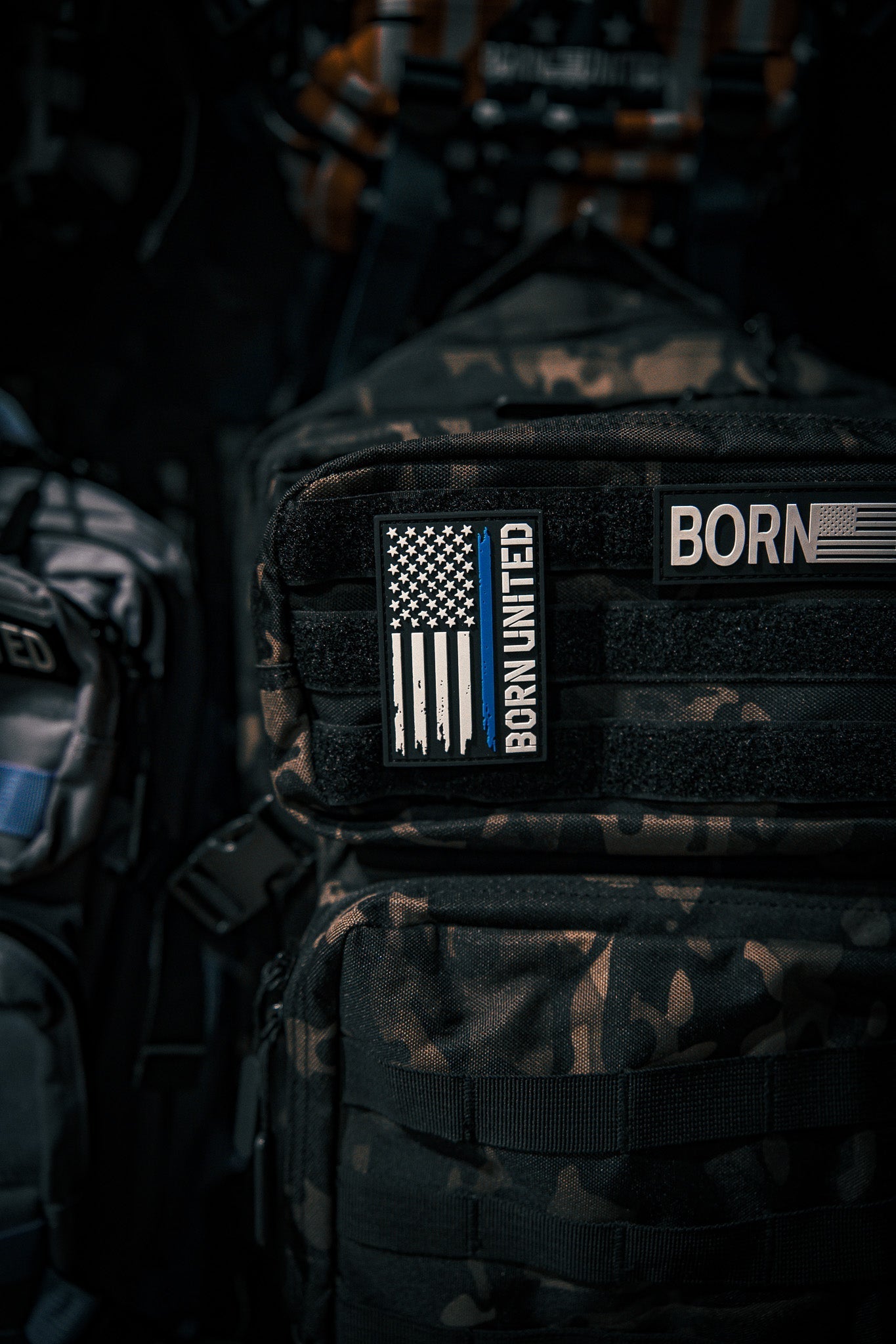 A camouflage backpack displays the Born United Private Label Blue Line Flag PVC Patch featuring a black-and-white American flag with a blue stripe. More backpacks and gear appear in the background.