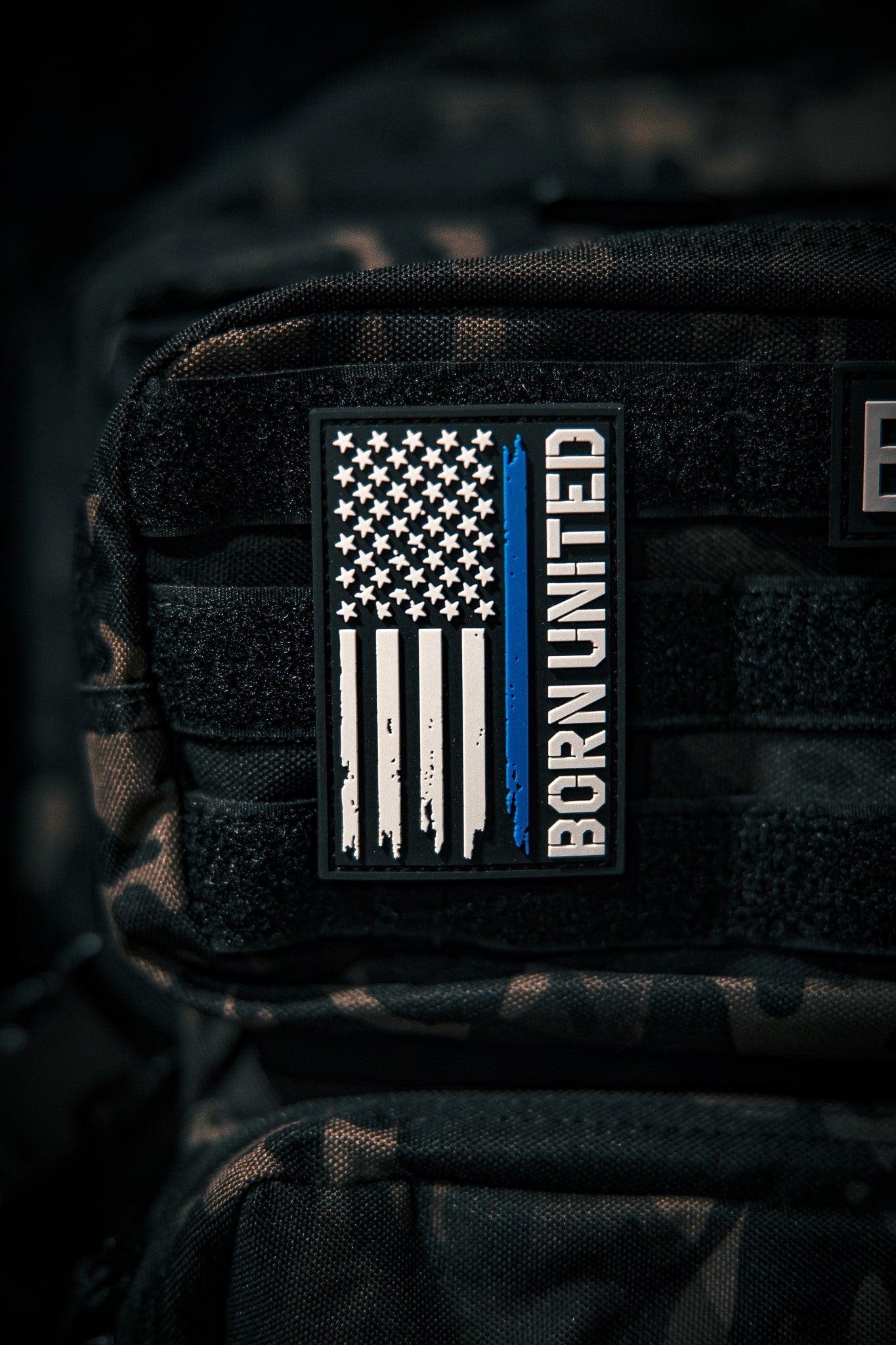 A Blue Line Flag PVC Patch by Born United Private Label, featuring a black, white, and blue American flag with a single blue stripe and BORN UNITED text, is attached to a black tactical backpack.