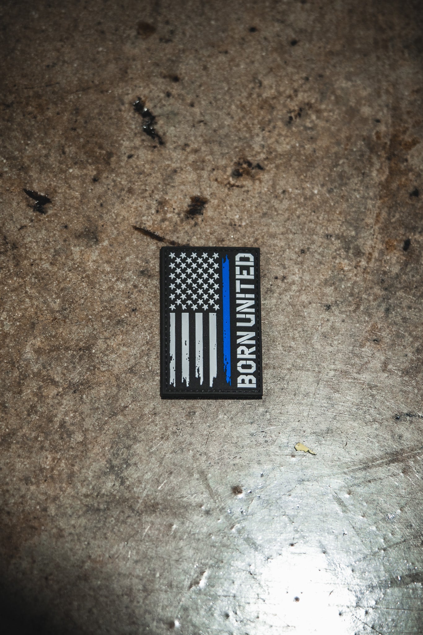 The Blue Line Flag PVC Patch by Born United Private Label features a black, white, and blue American flag with BORN UNITED written vertically on the right side, displayed on a textured surface.