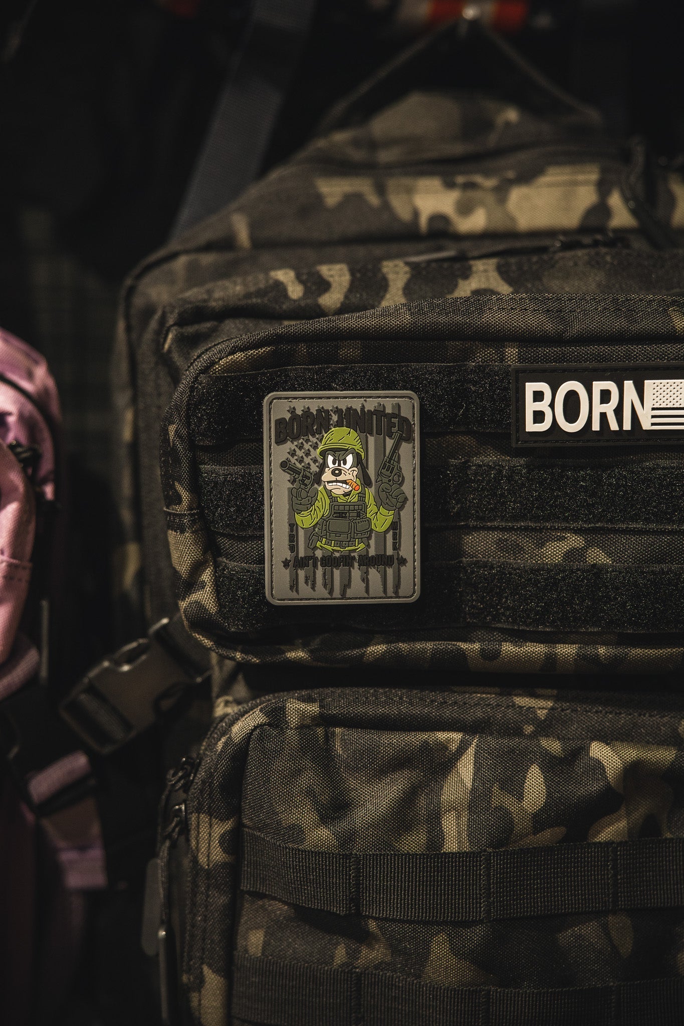 A camouflage backpack features a Born United Private Label Aint Goofin Around PVC patch showing a skeleton in military gear with rifles, attached to the front zip pocket.