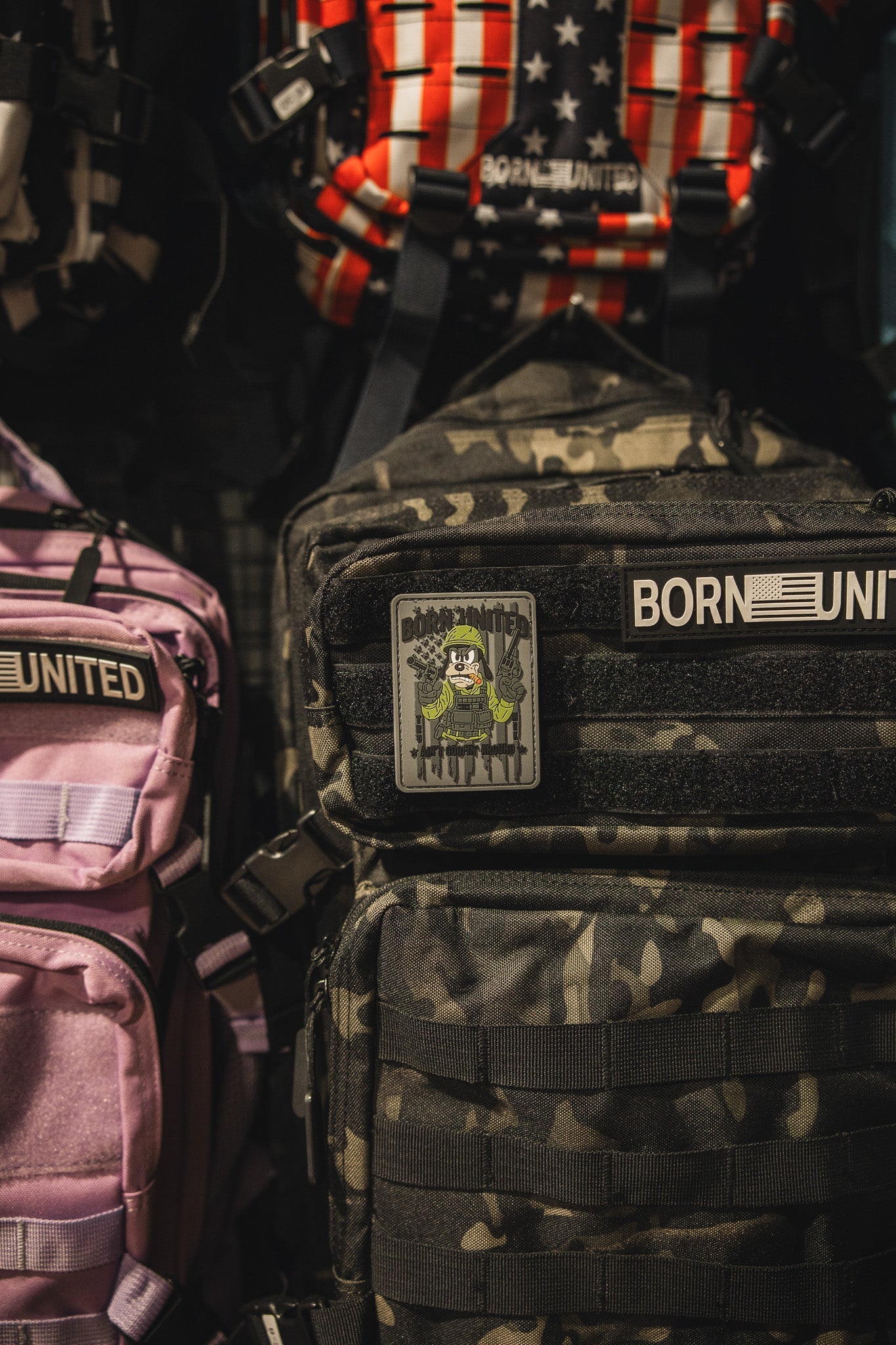 A close-up of several backpacks, including a black camo backpack with an Aint Goofin Around PVC Patch by Born United Private Label. A purple backpack is partially visible on the left.