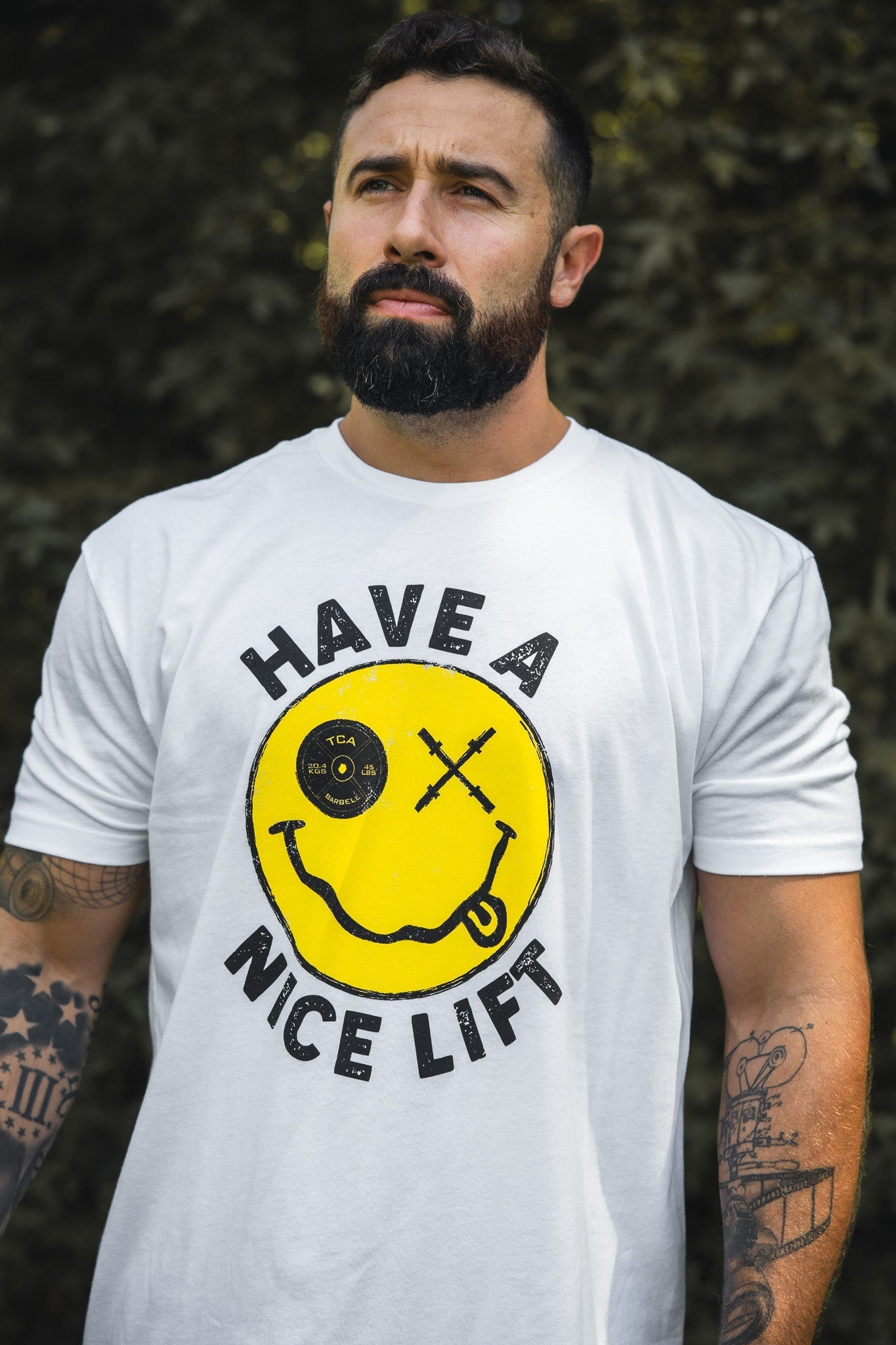 A bearded, tattooed man stands outdoors in greenery, wearing Tango Charlie Apparel’s “Thank You, have a Nice Lift” tee—a comfy cotton-poly blend shirt featuring a yellow smiley face and crossed barbells graphic.