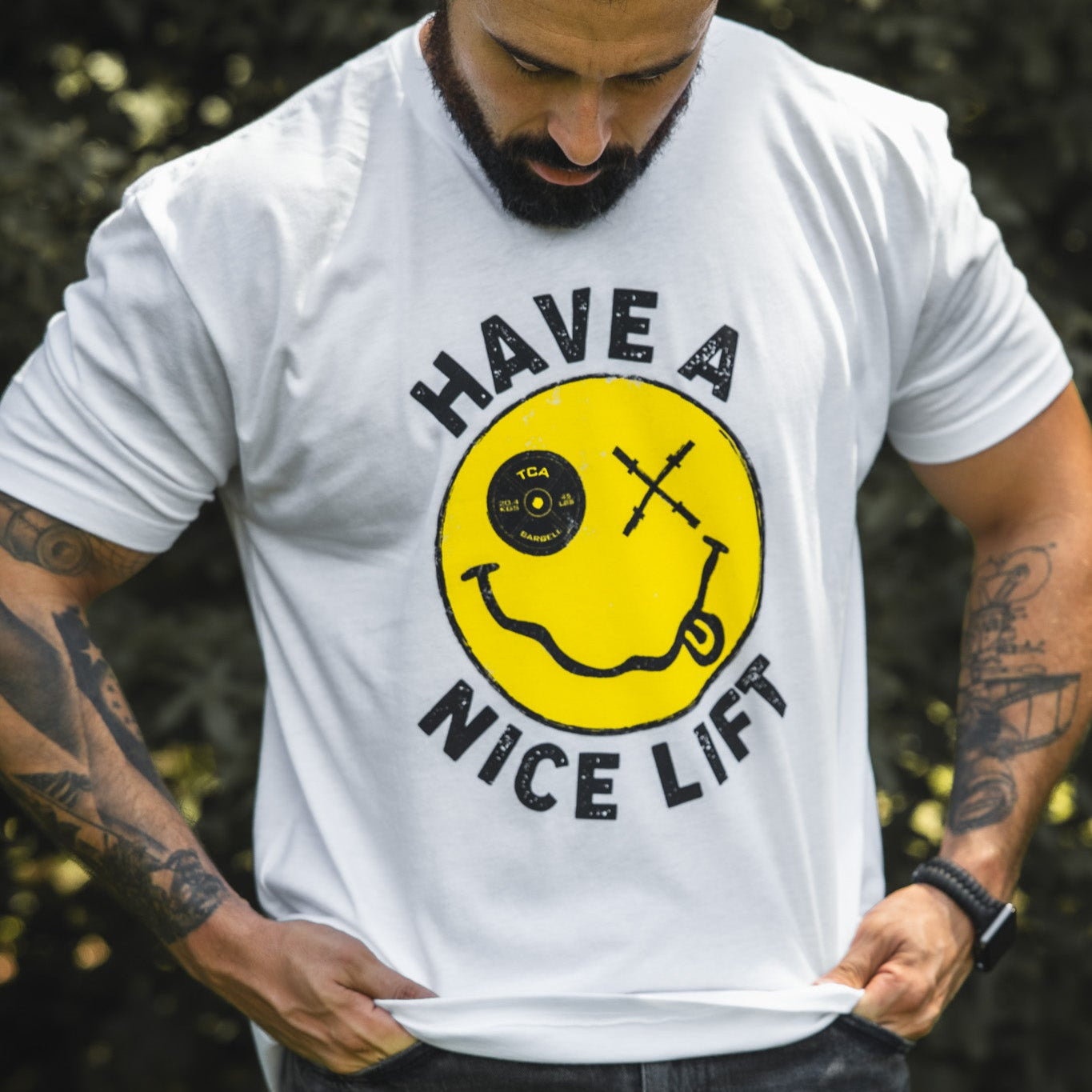A bearded man looks down at his comfy “Thank You, have a Nice Lift" tee by Tango Charlie Apparel, a white cotton-poly blend with a yellow smiley face featuring a bent barbell mouth, crossed barbell eyes, and “HAVE A NICE LIFT” text.