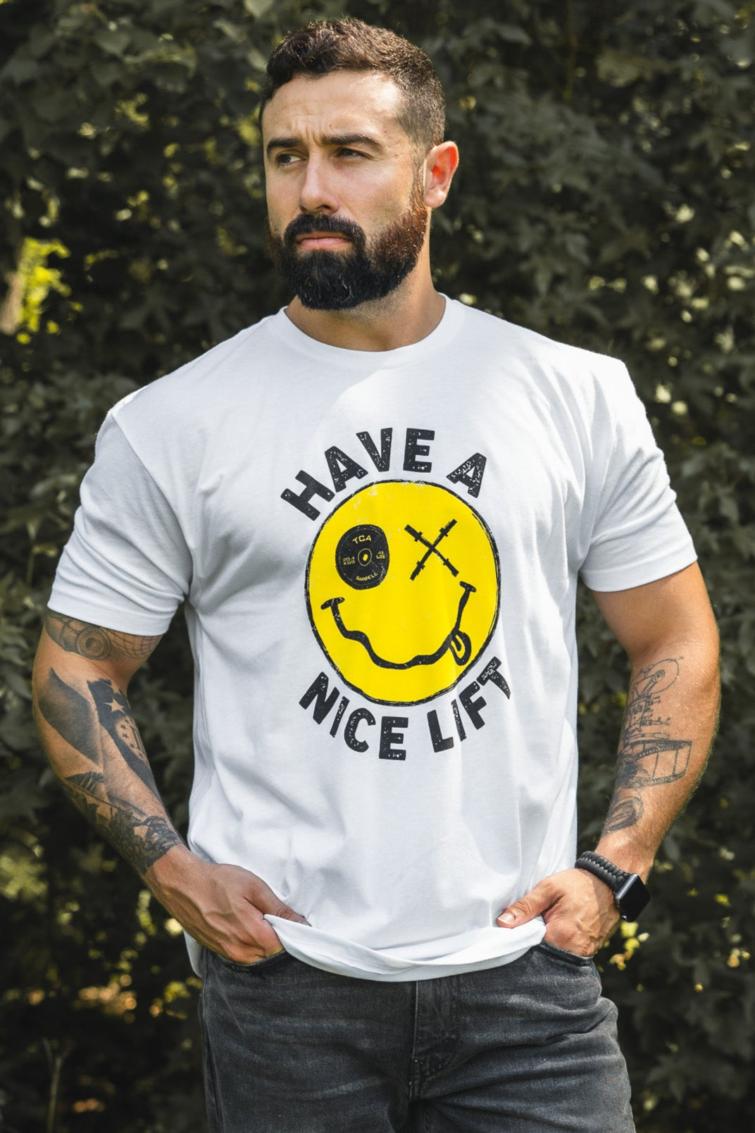 A tattooed, bearded man outdoors wears the Tango Charlie Apparel “Thank You, have a Nice Lift” tee—a comfy white cotton-poly shirt with a yellow smiley face lifting a barbell and the words HAVE A NICE LIFT.