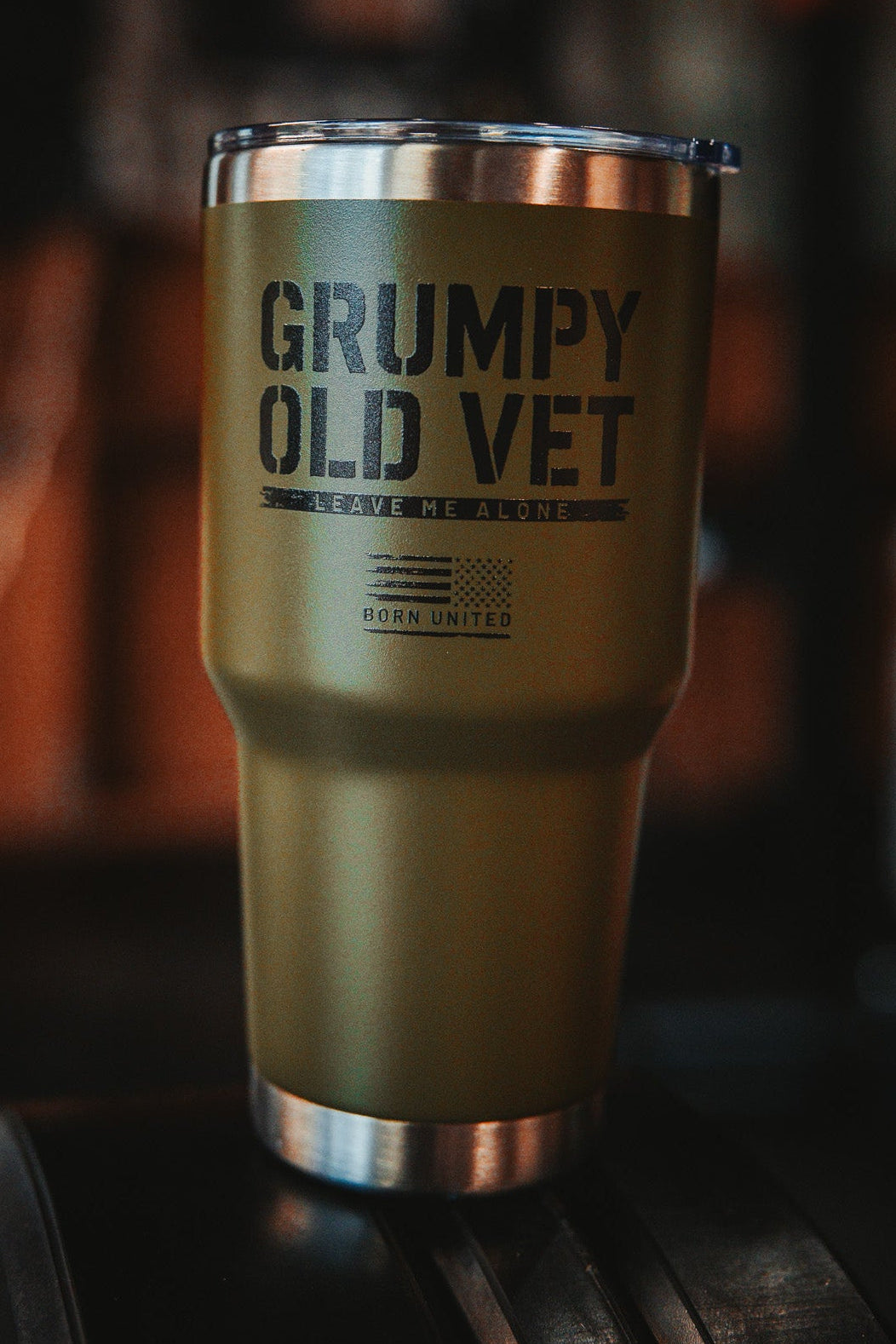 The Grumpy Old Vet Tumbler - 30oz from Born United Private Label, in green with a metal rim, displays bold black text: GRUMPY OLD VET, LEAVE ME ALONE, BORN UNITED against a blurred dark background.