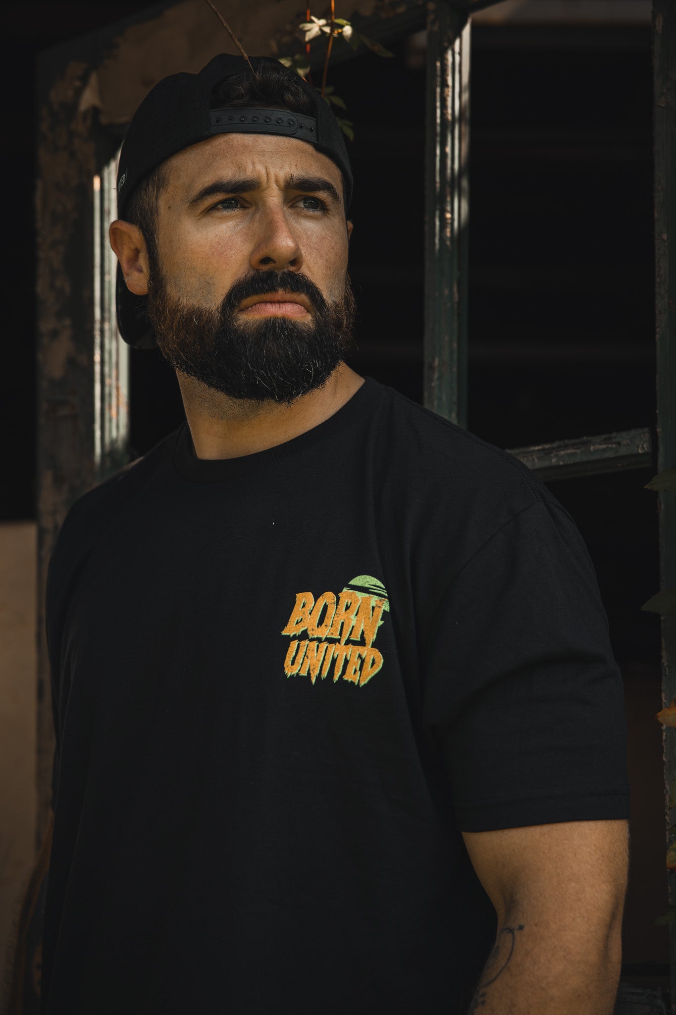 A bearded man in a black backwards cap and a Born United Private Label "Tankenstein" tee stands by a window, looking serious as he gazes into the distance.