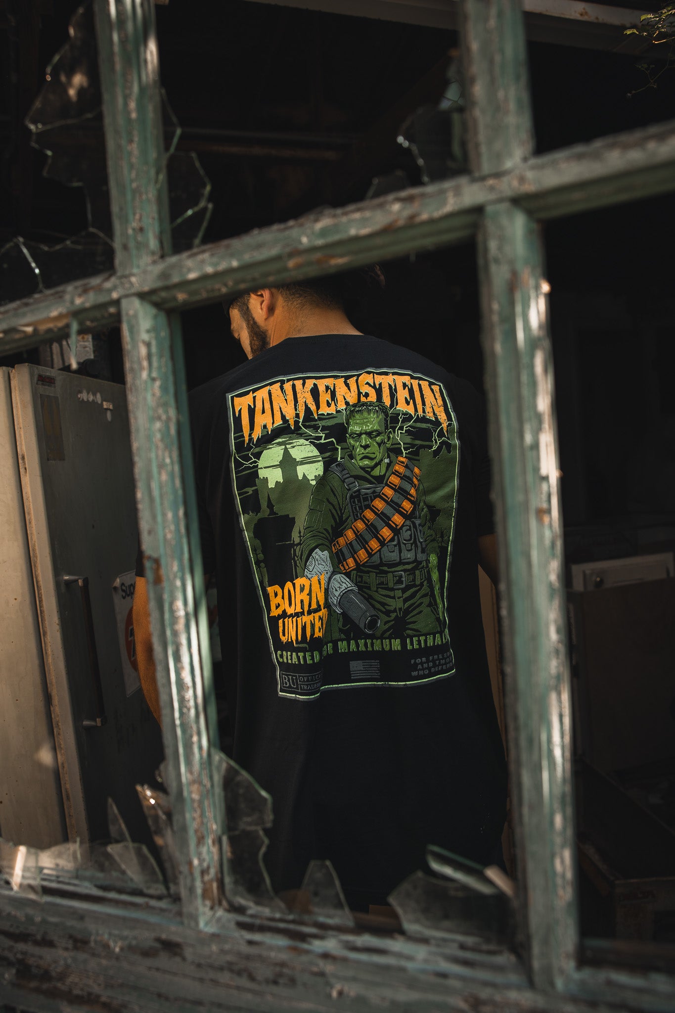 A man stands in a dim, abandoned building, seen through a broken window. He wears the limited edition Born United Private Label “Tankenstein” tactical tee featuring a green monster graphic.
