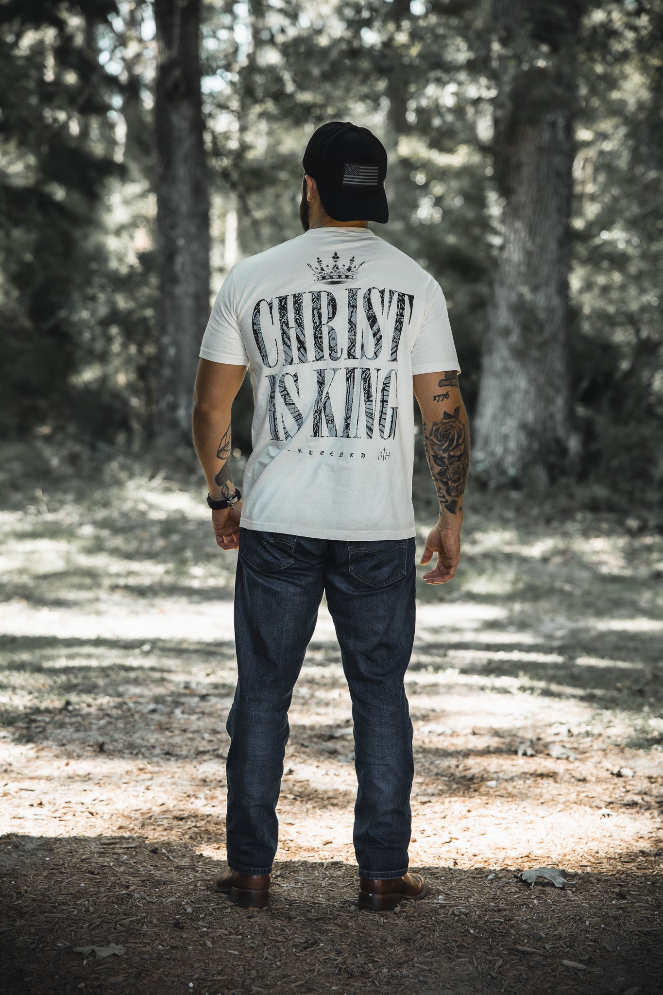 A man stands on a forest path, facing away, wearing the 13th Christ is King Premium Tee in Ivory with bold black lettering, blue jeans, a black cap, and tattoos on both arms.